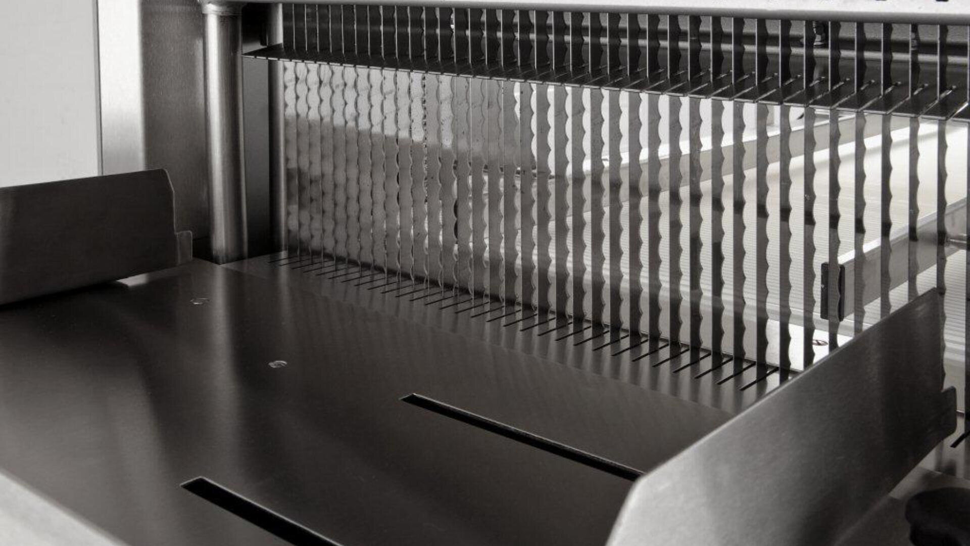 Nahaufnahme einer industriellen Lebensmittelschneidemaschine mit mehreren gleichmäßig angeordneten, gewellten Klingen, die zum Schneiden von Produkten wie Brot oder Käse in gleichmäßige Scheiben verwendet wird. Das Gerät besteht aus Metall und hat eine saubere, polierte Oberfläche.