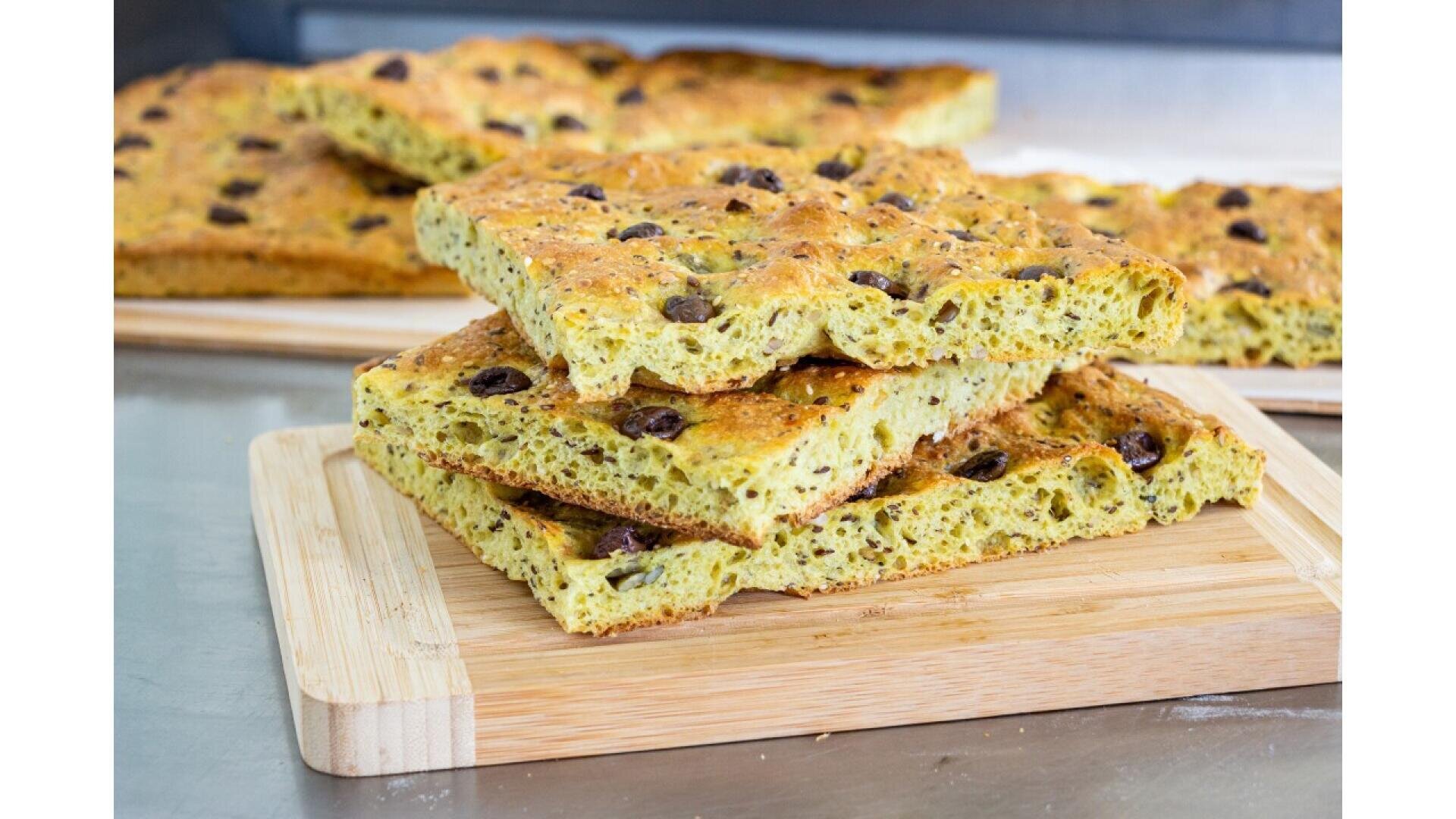 Drei rechteckige Stücke Oliven-Focaccia, die auf einem hölzernen Schneidebrett gestapelt sind, mit weiteren Focaccia-Stücken im Hintergrund auf einer Küchenzeile. Das Brot hat eine goldene Kruste und sichtbare Oliven.