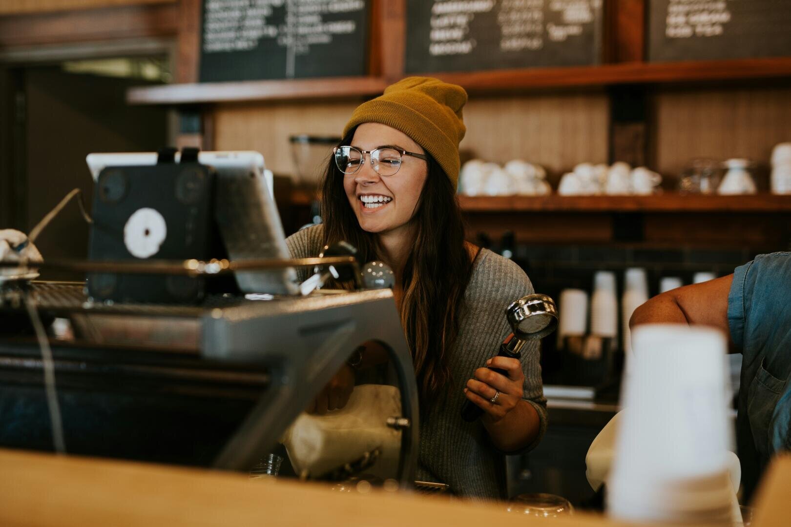 Ein lächelnder Barista mit Beanie und Brille steht hinter einer Kaffeetheke, hält einen Siebträger und interagiert mit einer Touchscreen-Kasse in einem gemütlichen Café-Ambiente.