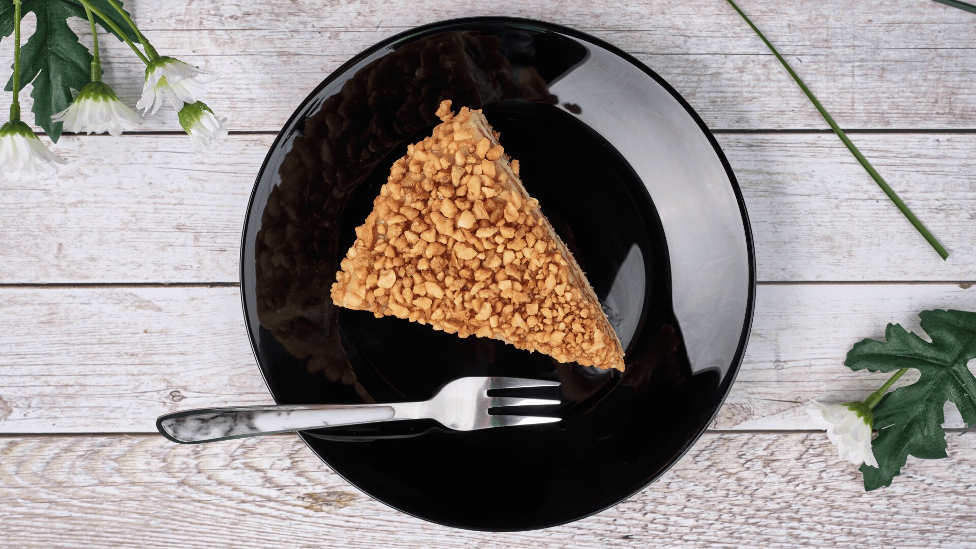 A slice of cake with a nut topping on a black plate, accompanied by a fork, sits on a white wooden surface with white flowers nearby.