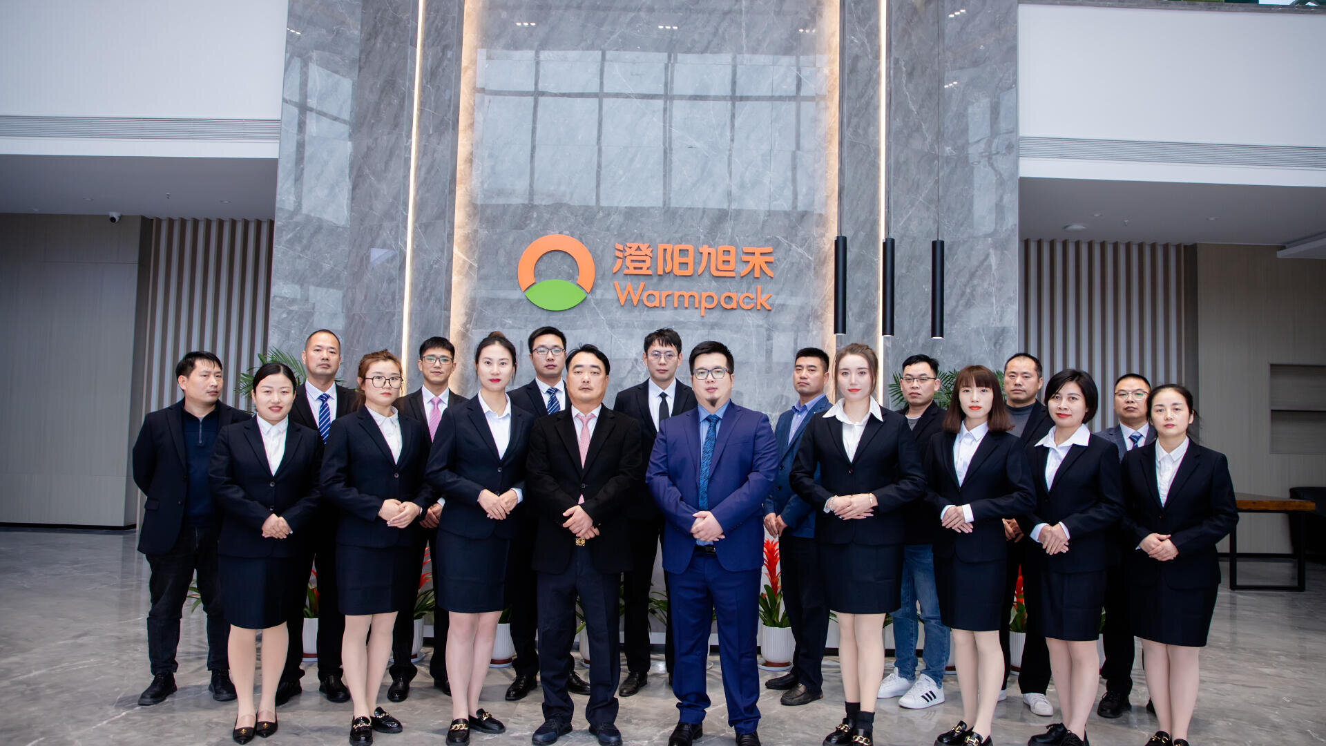 A group of professionals in formal attire stand in rows in front of a marble wall with the Warmpack logo and name, posing for a team photo in a modern office lobby.