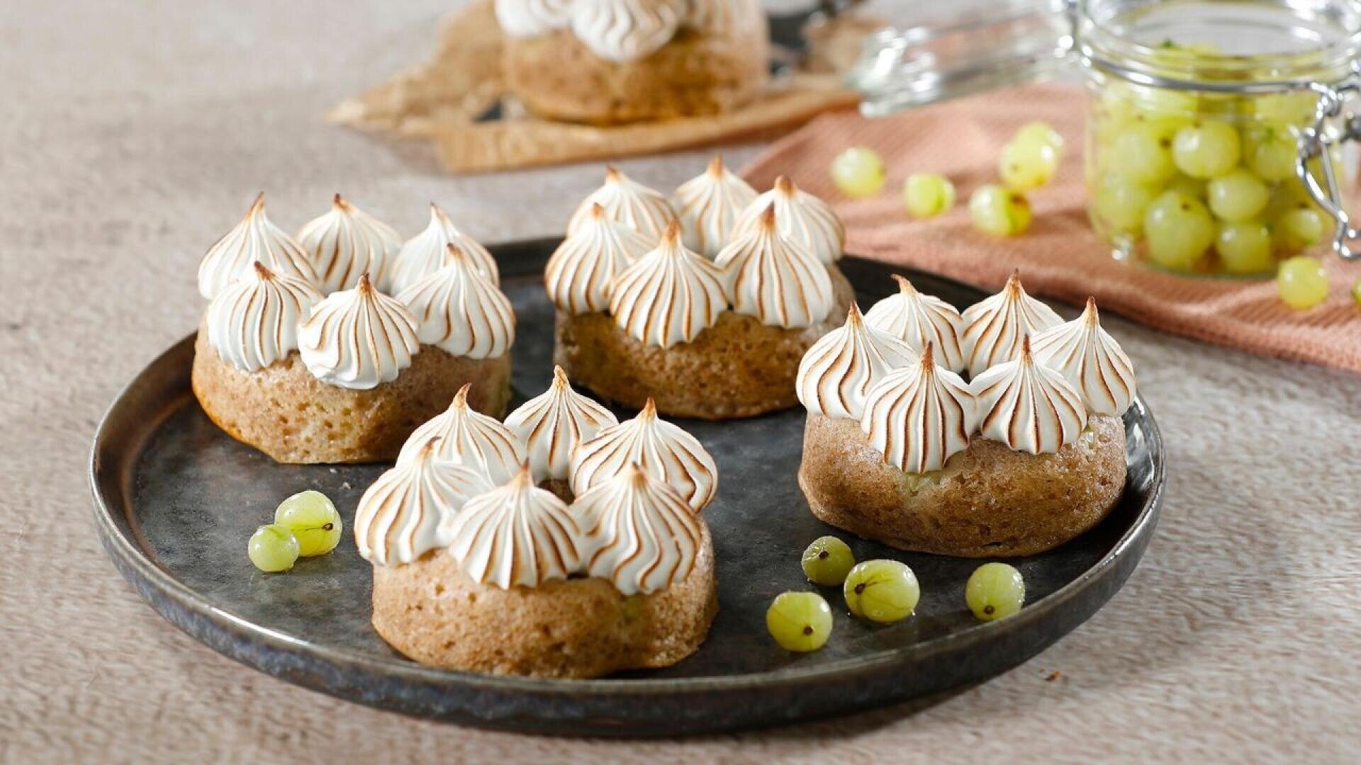 A dark plate with four small cakes topped with piped toasted meringue peaks. Green gooseberries are scattered around, with more in a glass jar and on a cloth in the background.