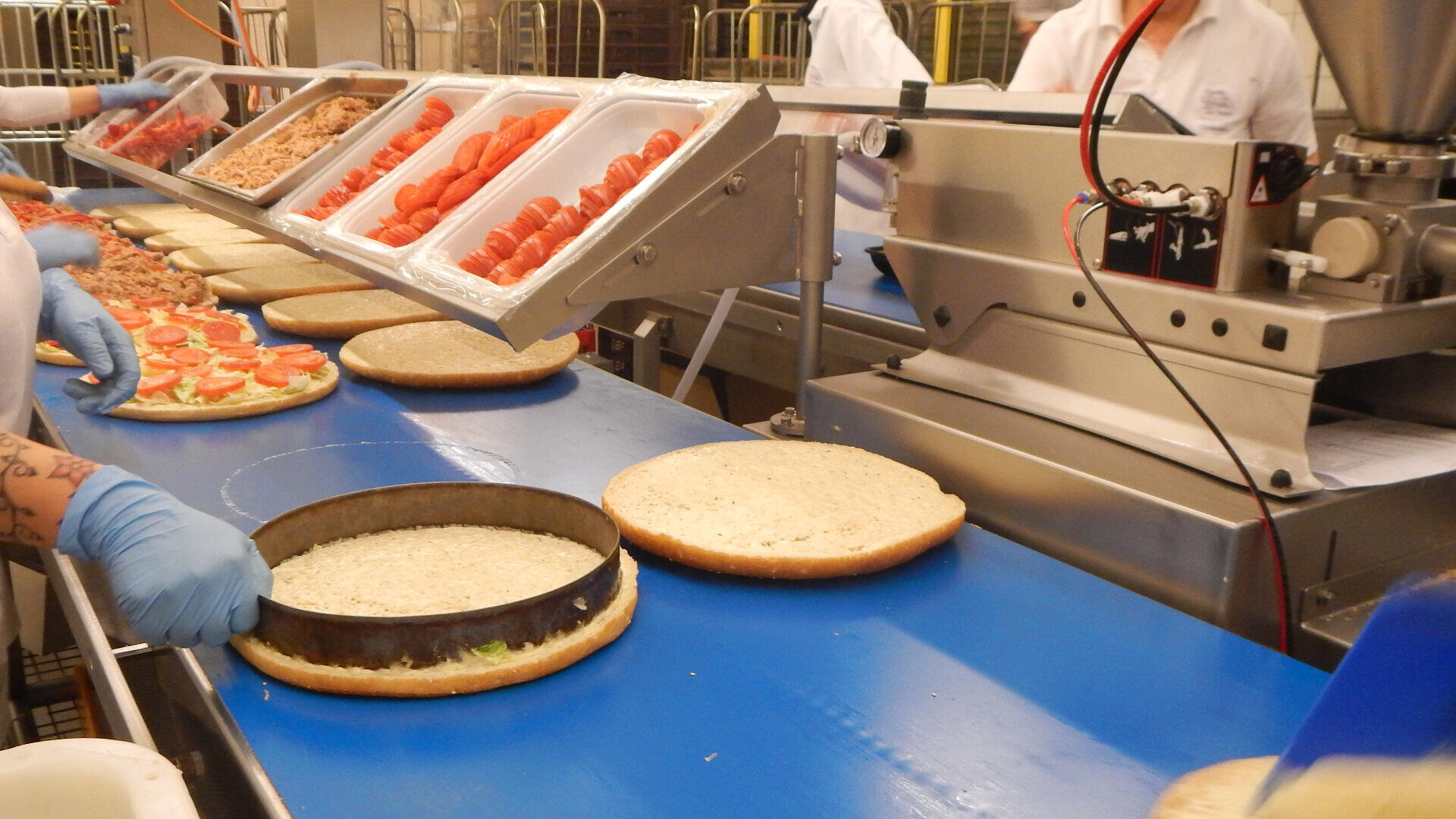 Arbeiter mit Handschuhen montieren Sandwiches oder Burger auf einem blauen Fließband in einer Lebensmittelverarbeitungsanlage, wobei Schalen mit geschnittenen Tomaten und anderen Zutaten sichtbar sind.