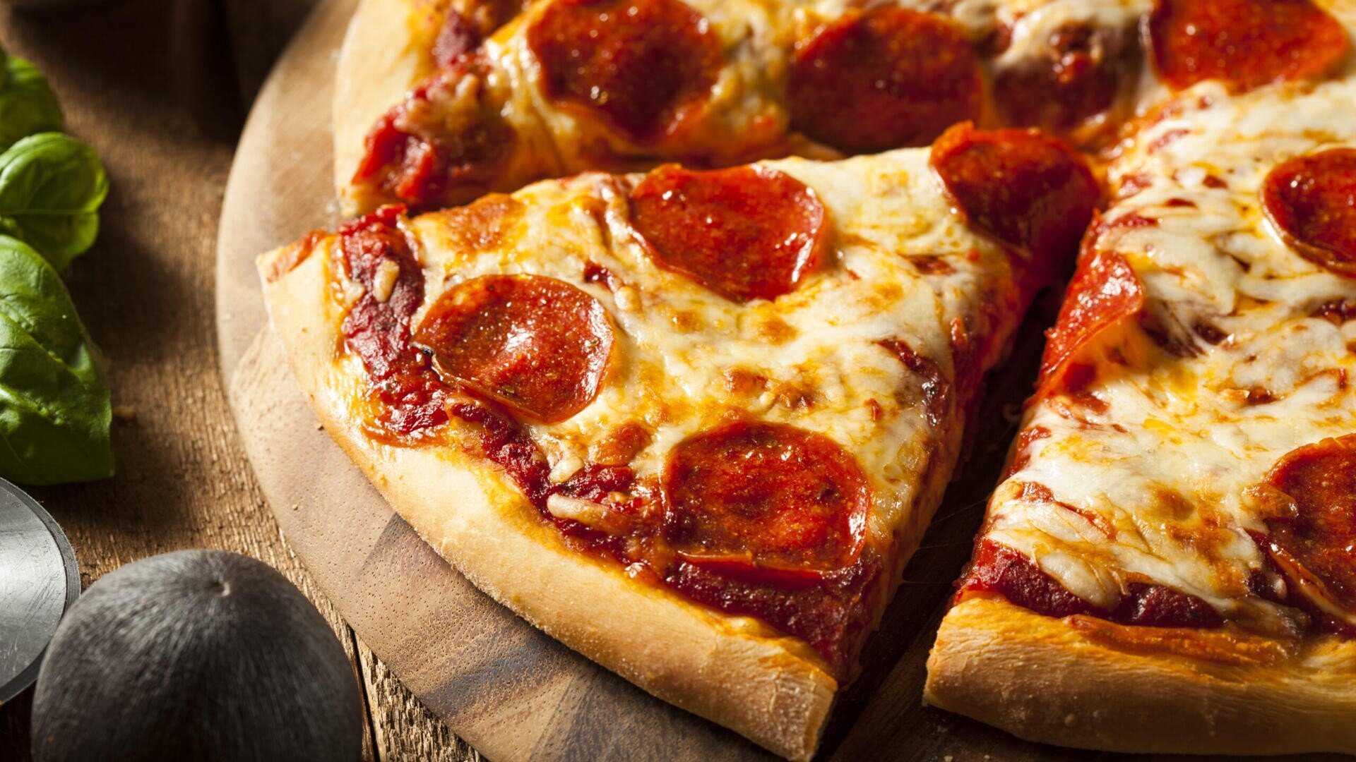 Nahaufnahme von Peperoni-Pizzastücken auf einem Holzbrett, belegt mit geschmolzenem Käse und Peperoni, mit einem Teil eines frischen Basilikumzweigs und einem runden dunklen Objekt am Rand.