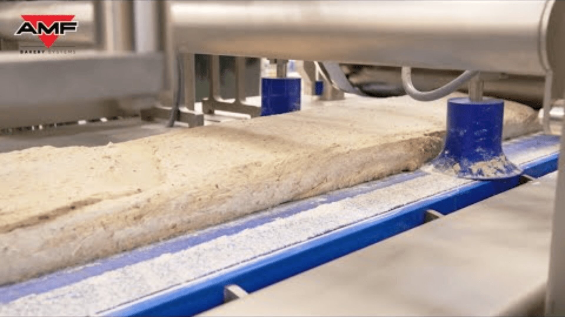A close-up of an industrial dough processing machine shaping a large slab of dough on a conveyor belt, with the AMF company logo visible in the upper left corner.