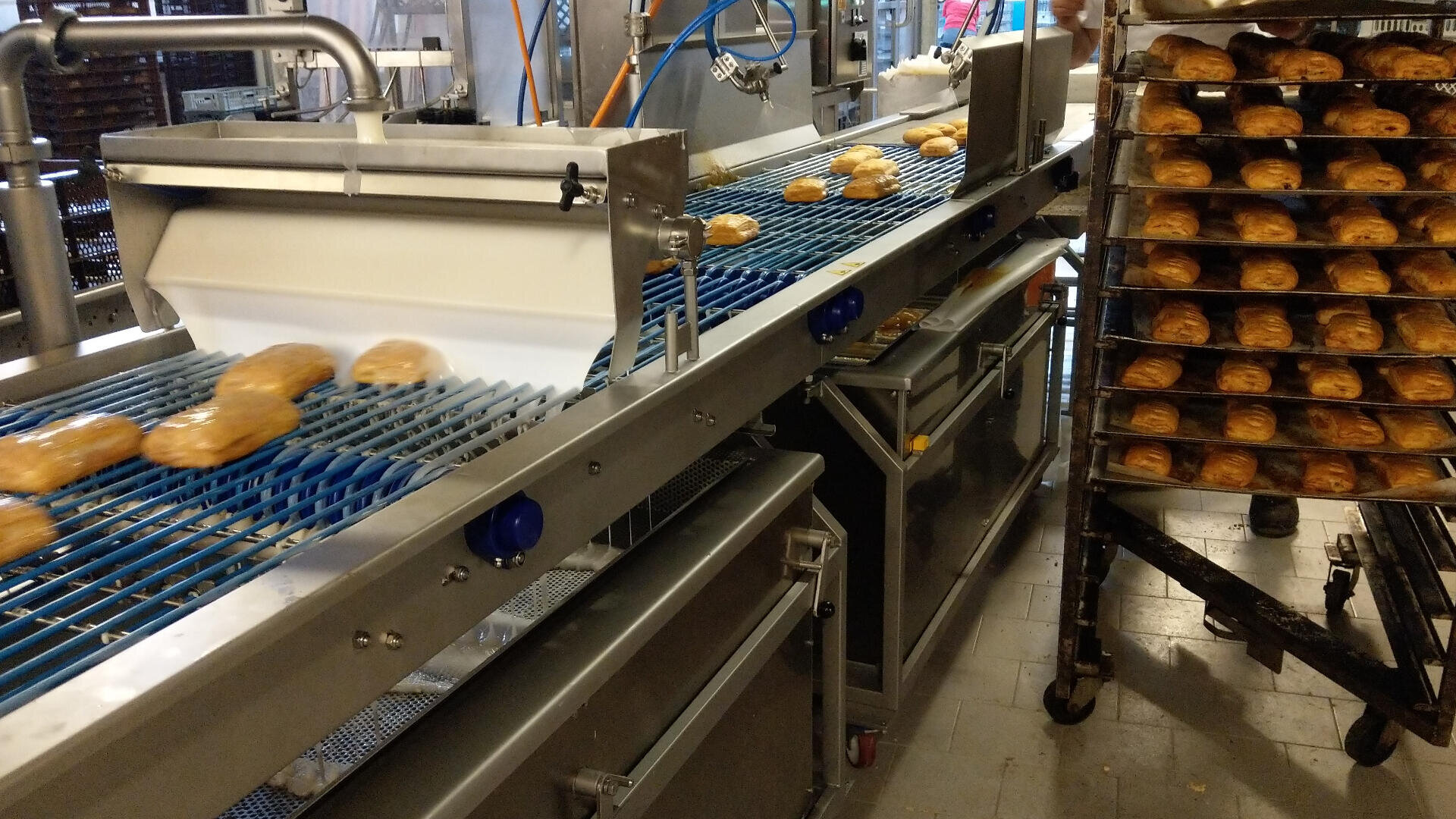 Pastries move along a conveyor belt in a large industrial bakery, with trays of baked goods stacked on a cart nearby. Machinery and equipment are visible in the background.