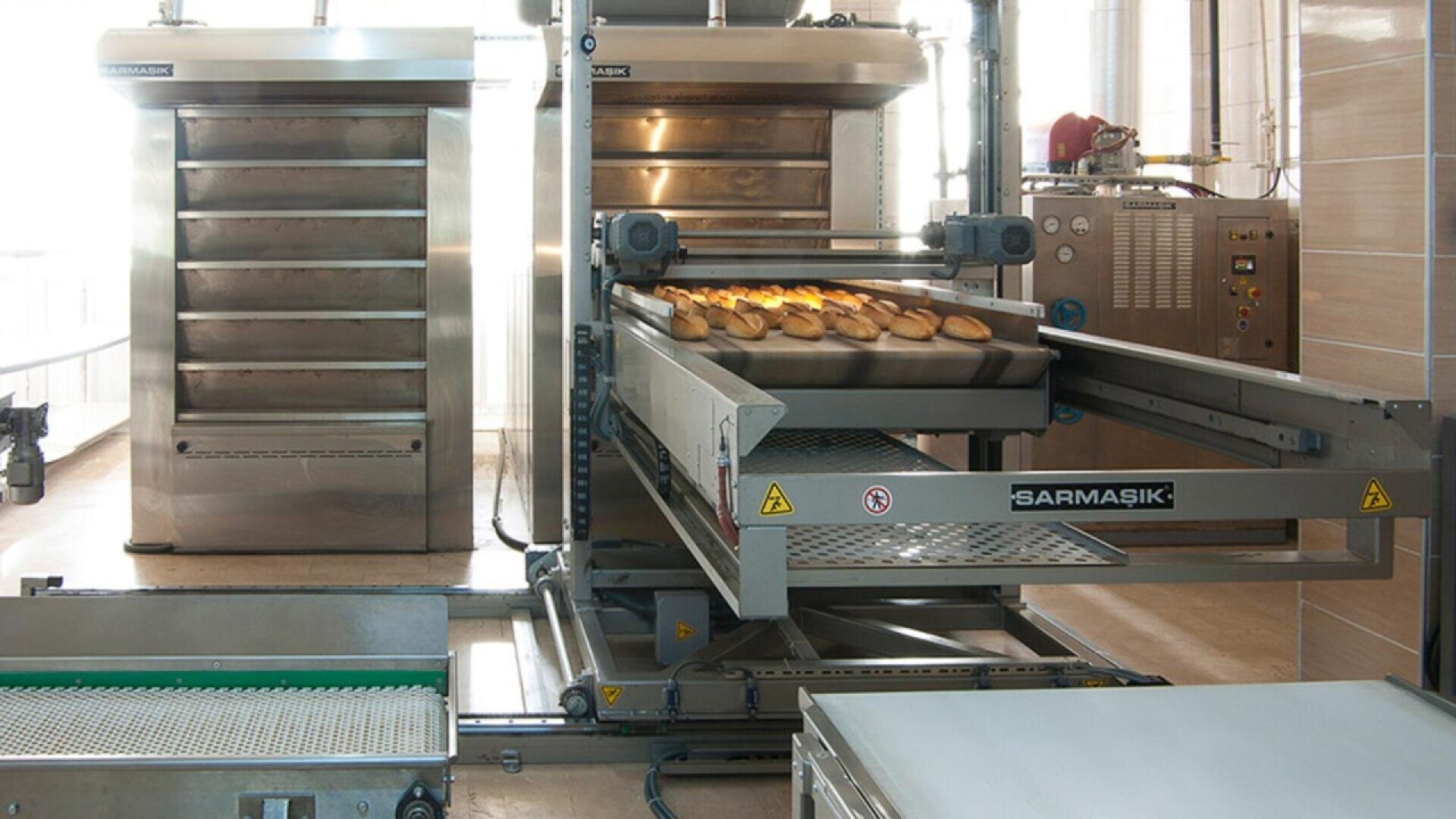 A commercial bread oven with multiple trays and a conveyor belt is baking several loaves of bread in an industrial bakery setting. Stainless steel equipment surrounds the bread as it moves out of the oven.