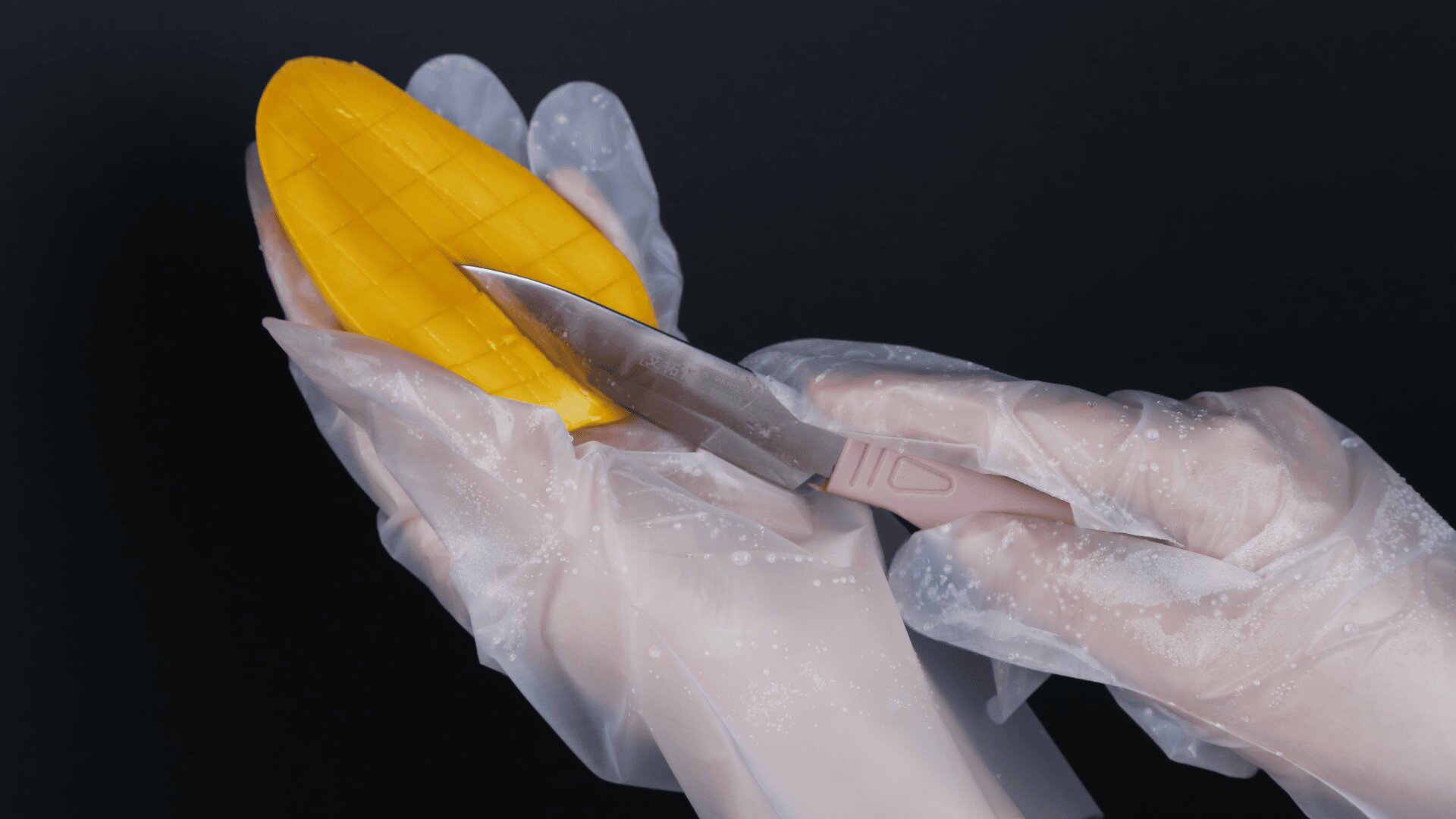 A person wearing translucent gloves holds half a mango while cutting a grid pattern into the fruit with a knife against a dark background.