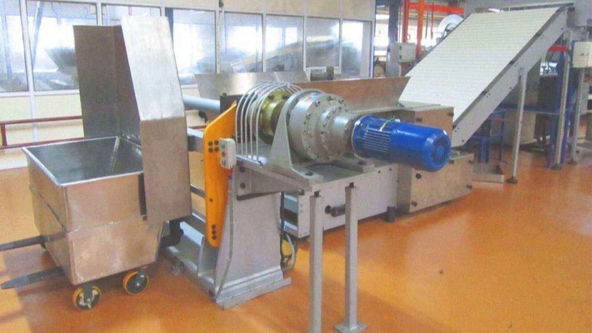 A stainless steel industrial food processing machine with a large blue motor, conveyor belt, and collection container in a clean factory setting with orange flooring and glass-paneled walls.