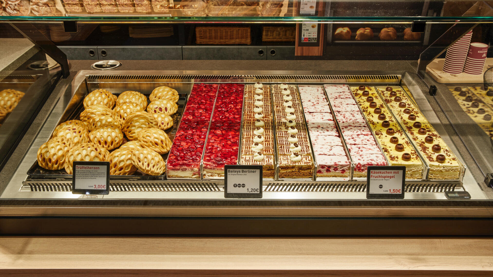 Eine Bäckereivitrine mit drei Arten von Gebäck: links gitterförmige Apfeltaschen, in der Mitte mit roten Beeren belegte Scheiben und rechts Sahnetorten mit Kirschbelag, jeweils mit Preisschildern versehen.
