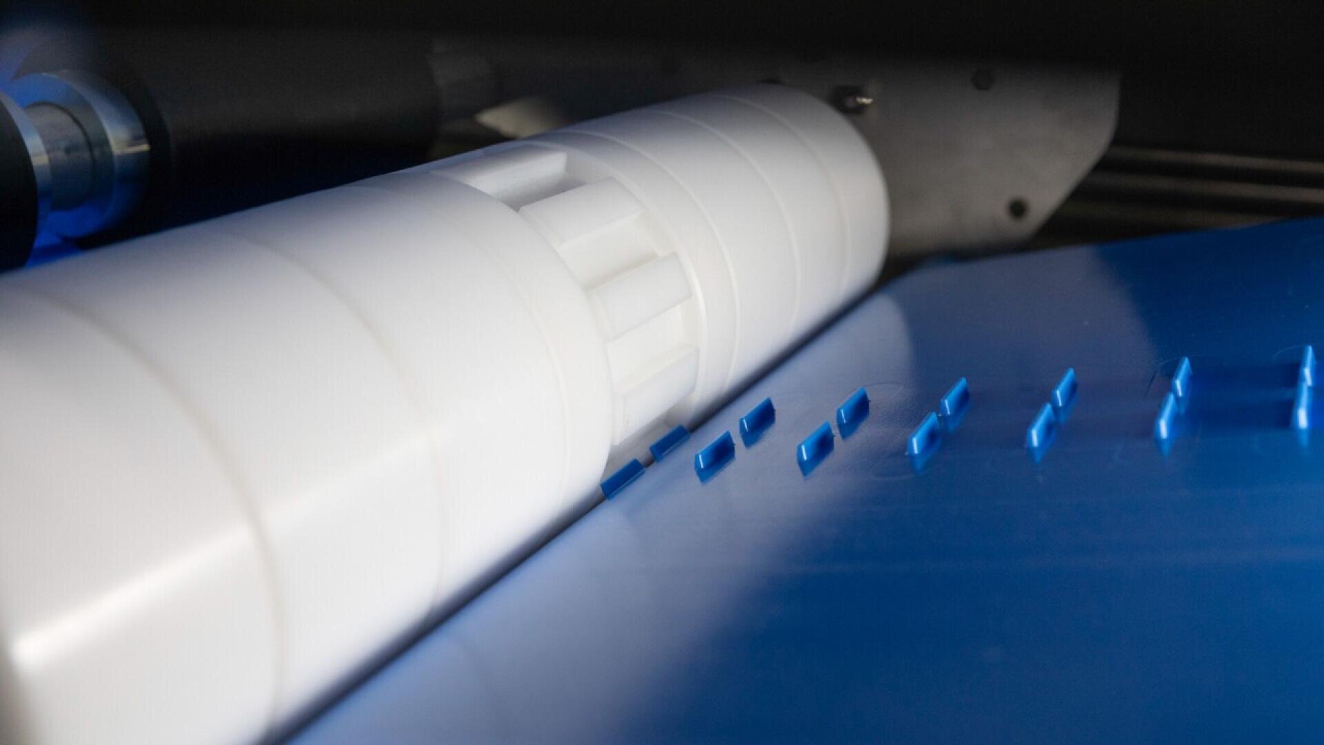 Nahaufnahme eines weißen zylindrischen Zahnradmechanismus, der mit einer blauen Kunststoffplatte mit rechteckigen Ausschnitten interagiert, was auf einen industriellen oder Herstellungsprozess hindeutet.