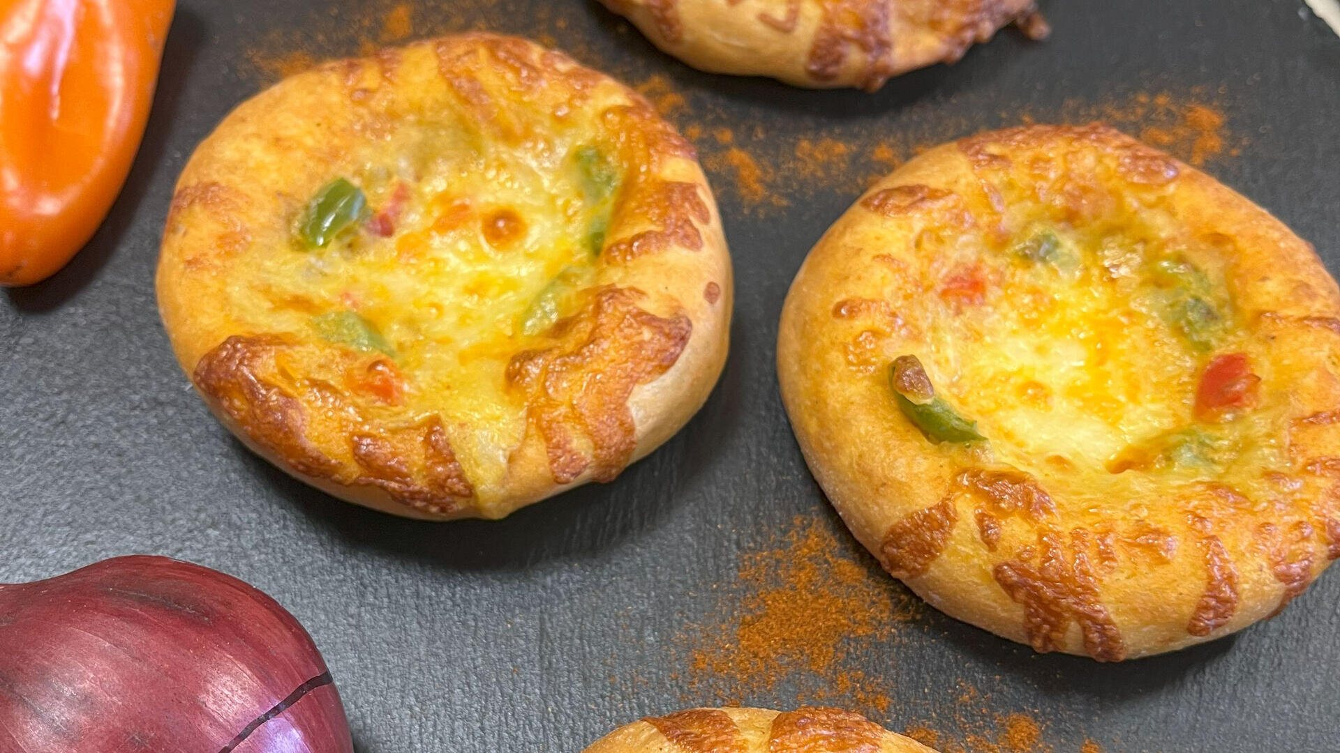Mini-Käse- und Gemüsebrotschalen auf schwarzem Untergrund, umgeben von Paprikapulver und einem Teil einer roten Zwiebel und orangefarbener Paprika.