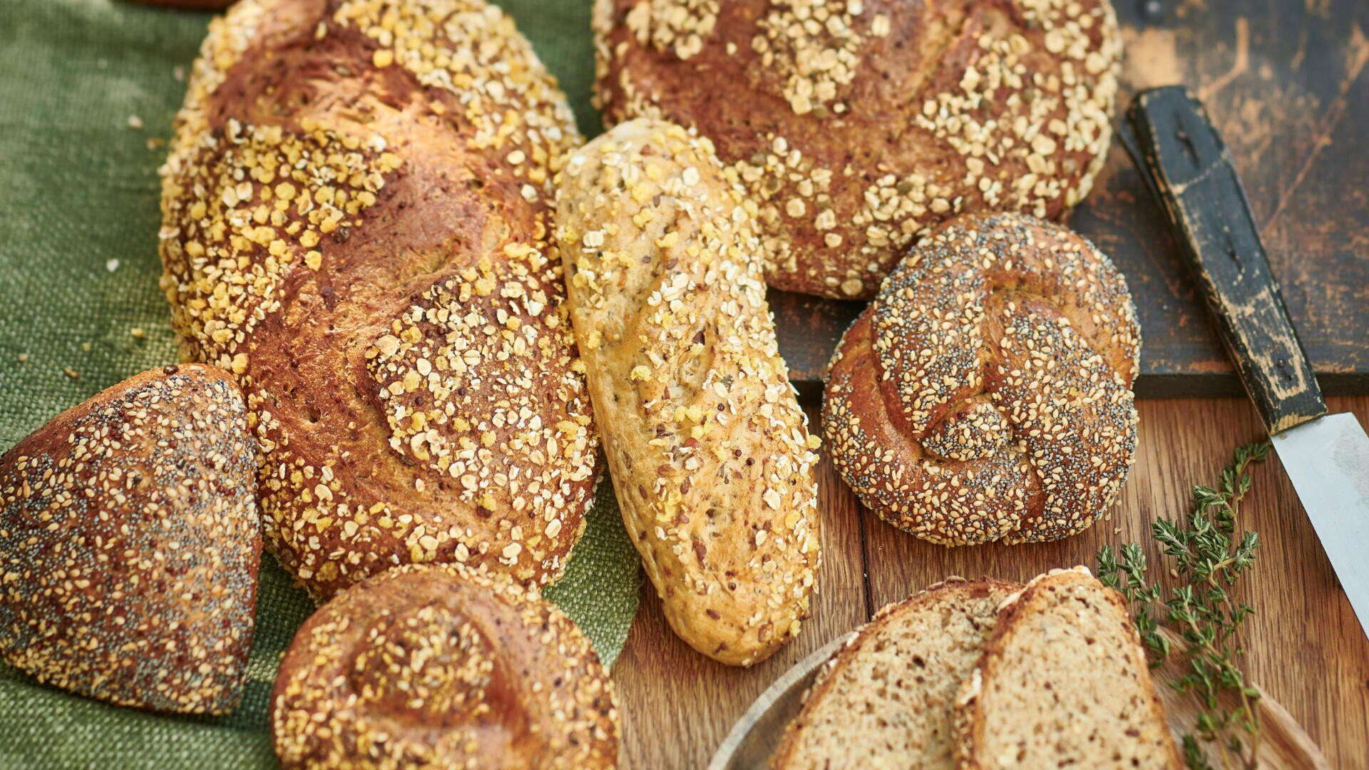 Verschiedene mit Körnern und Haferflocken belegte Vollkornbrote, darunter Brötchen und Brotscheiben, liegen auf einer Holzfläche mit einem grünen Tuch und einem Brotmesser in der Nähe.