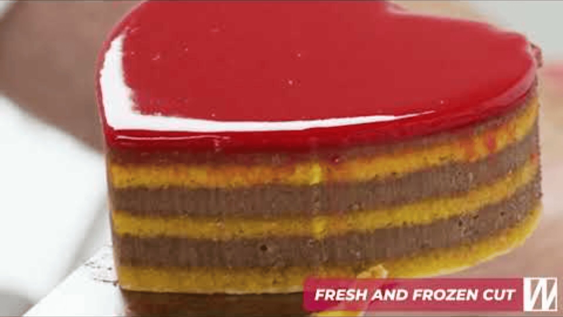 A heart-shaped layered dessert with glossy red glaze on top and alternating yellow and brown layers, shown being held on a spatula. A label reads “FRESH AND FROZEN CUT” in the corner.