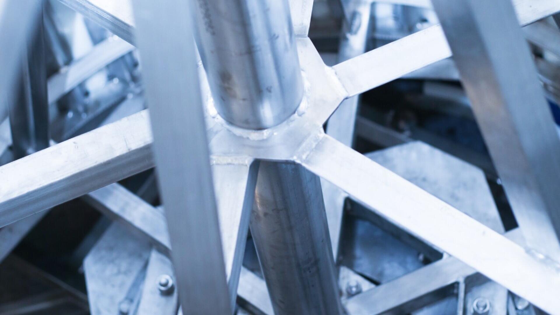 Nahaufnahme sich kreuzender Metallträger und -stangen, die eine geometrische, industrielle Struktur in kühlem Blauton bilden. Das Bild hebt die klaren, kantigen Linien und glänzenden Oberflächen des Metallgerüsts hervor.