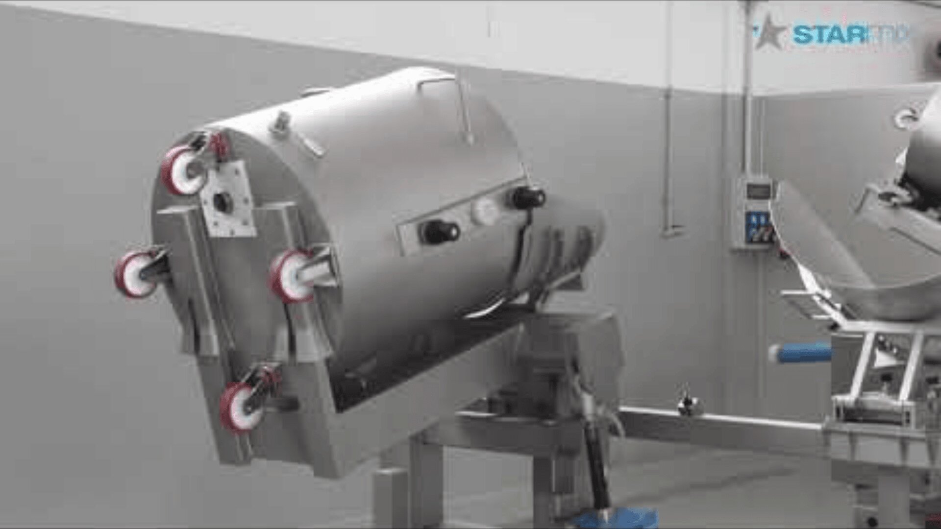 A large industrial stainless steel machine with red-and-white rollers is mounted on a metal frame in a gray, sterile room. Control panels and equipment can be seen in the background.