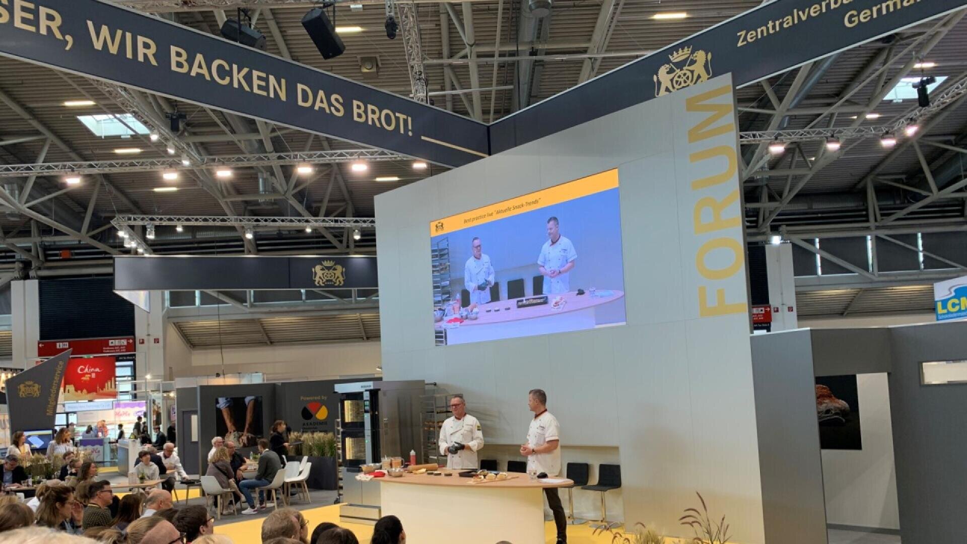 Two men give a baking demonstration on stage at a trade show, with a large screen displaying their actions. An audience is seated, and banners with German text and a bread association logo hang above.