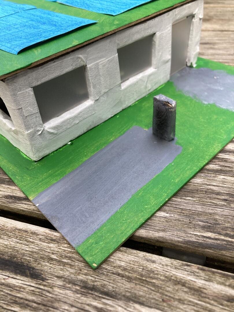 Ein handgefertigtes Modell eines weißen Gebäudes mit blauen Dachabschnitten, drei Fensteröffnungen und einer Auffahrt. In der Nähe der Auffahrt steht ein kleines zylindrisches Objekt, das auf einem grünen Sockel auf einer Holzoberfläche steht.