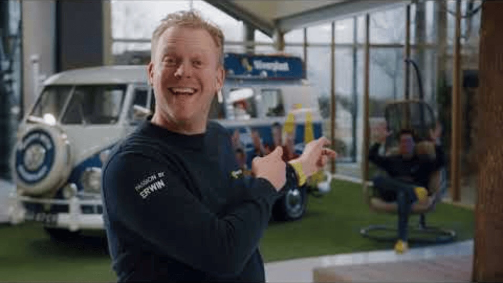 A smiling man points excitedly over his shoulder at another man sitting in a hanging chair, with a vintage van and bright indoor setting in the background.