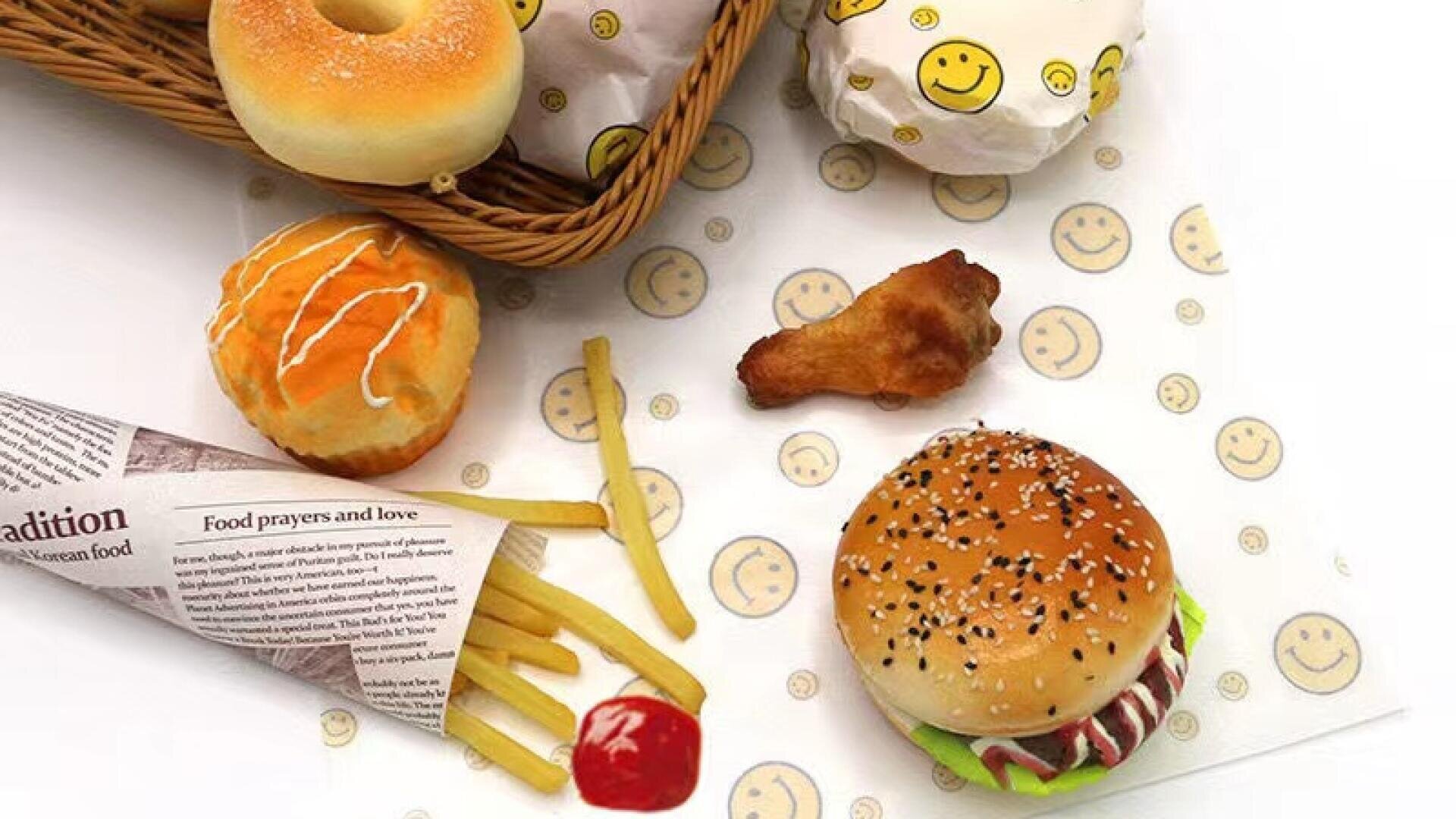 A sesame seed hamburger, fries with ketchup, a chicken wing, a donut, and a pastry are arranged on smiley face paper. Some items are wrapped or served in a paper basket.