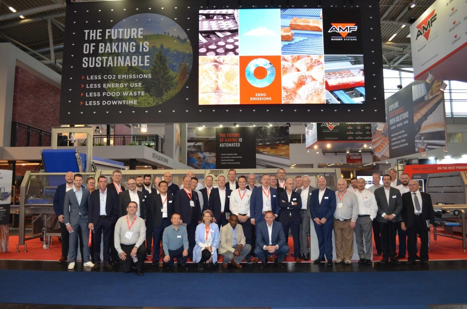 A group of around 40 people pose for a photo at a trade show booth for AMF Bakery Systems. Behind them are large signs promoting sustainable baking and a screen showing bakery images and environmental messaging.