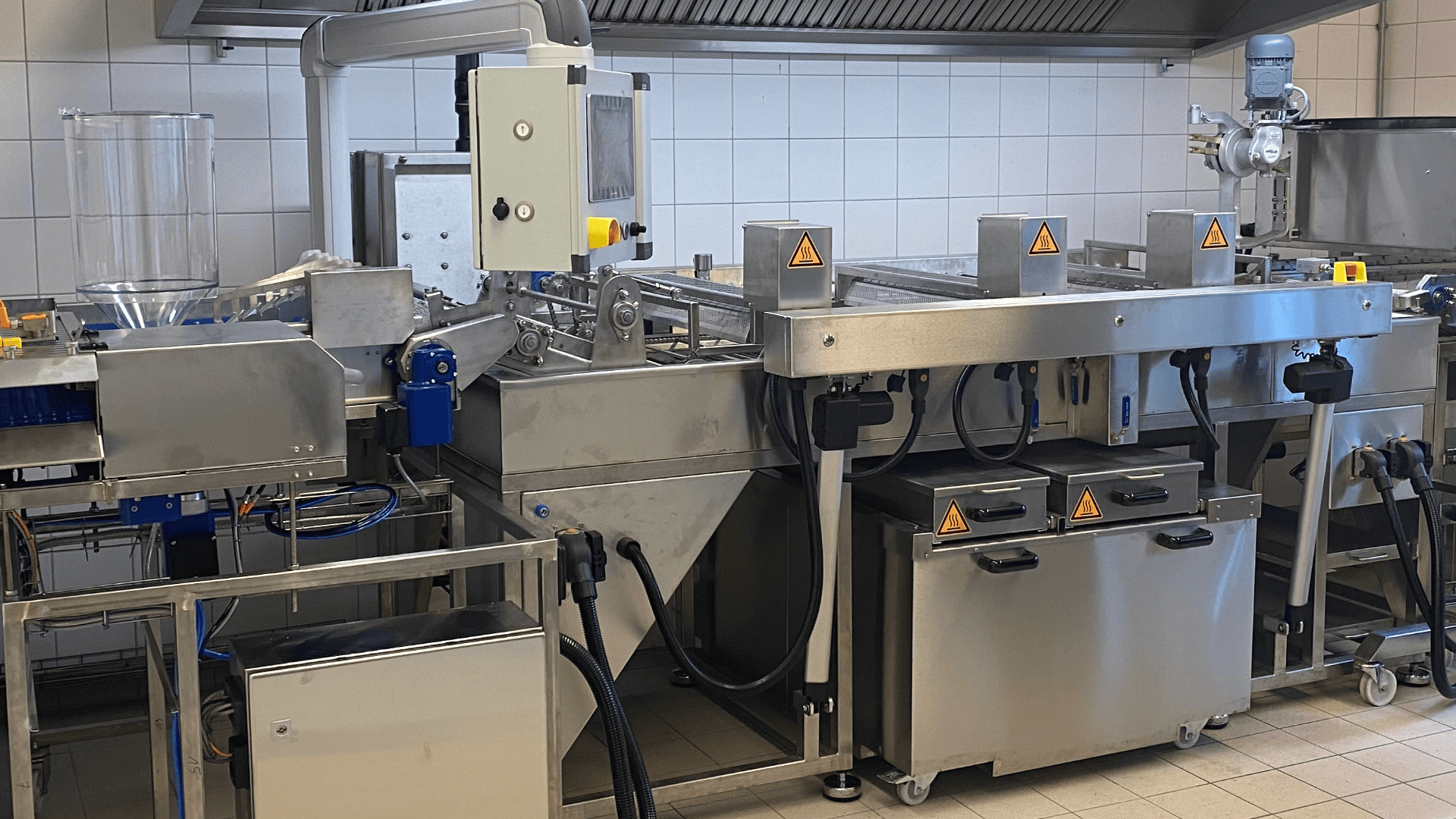 In einer gefliesten Großküche steht eine industrielle Lebensmittelverarbeitungsmaschine aus Edelstahl mit Förderbändern, Bedienfeldern, Warnschildern und verschiedenen miteinander verbundenen Fächern.