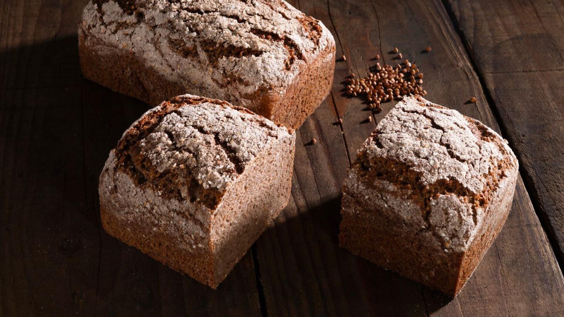 Drei Laibe dunkles, rustikales Brot mit einer rissigen, mit Mehl bestäubten Kruste liegen auf einer Holzoberfläche, daneben ist ein kleiner Haufen Leinsamen verstreut.