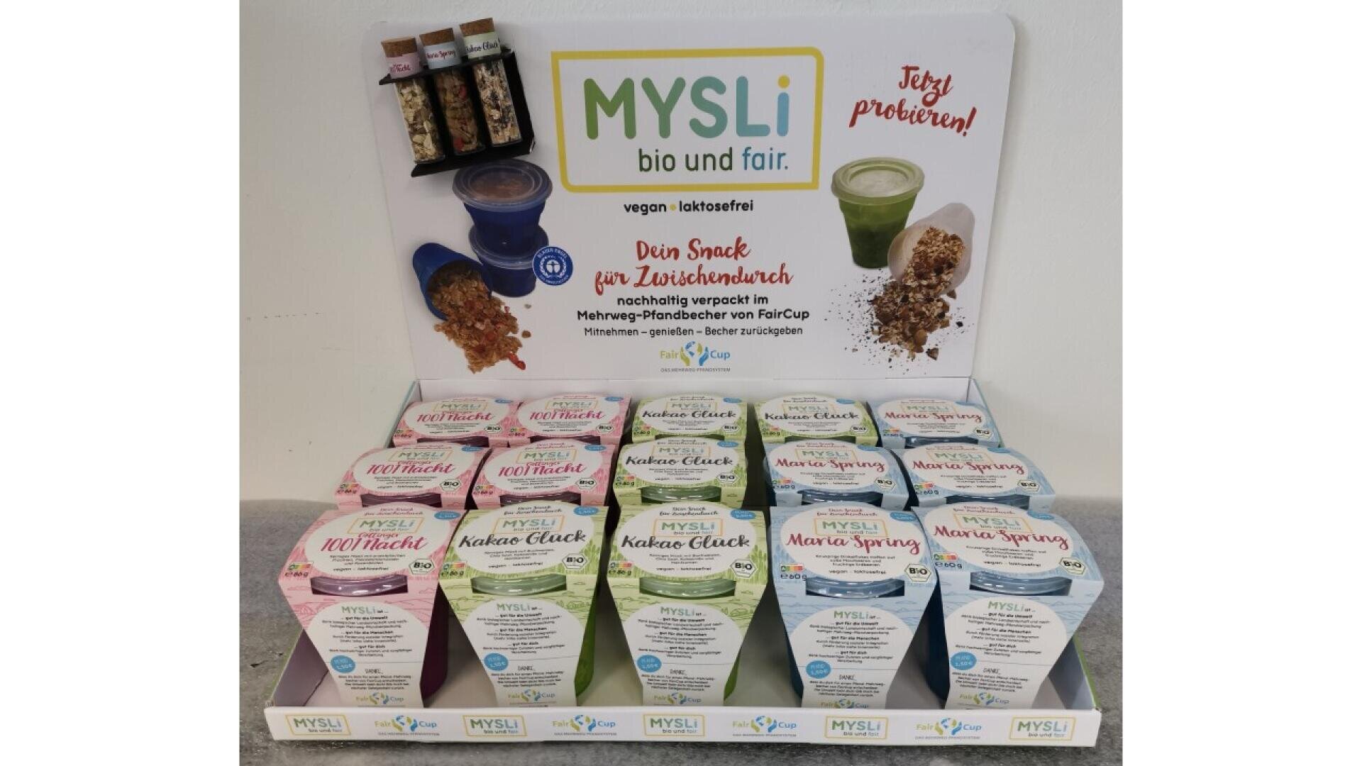 Display of MYSLI muesli snacks in colorful containers with labels indicating vegan and lactose-free options. The sign behind promotes sustainable, fair packaging and encourages customers to try the products.