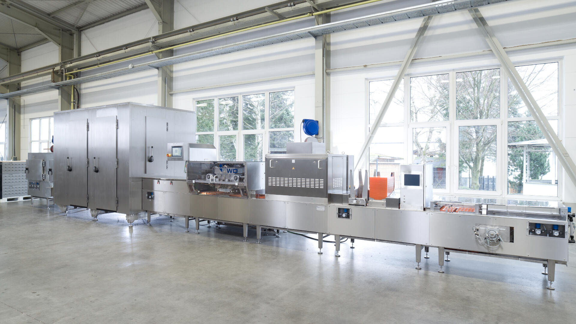 Eine moderne industrielle Lebensmittelverarbeitungsmaschine in einer sauberen, geräumigen Fabrik mit großen Fenstern, die natürliches Licht hereinlassen. Die Anlage ist aus rostfreiem Stahl gefertigt und besteht aus mehreren Teilen, die über ein Förderband miteinander verbunden sind.