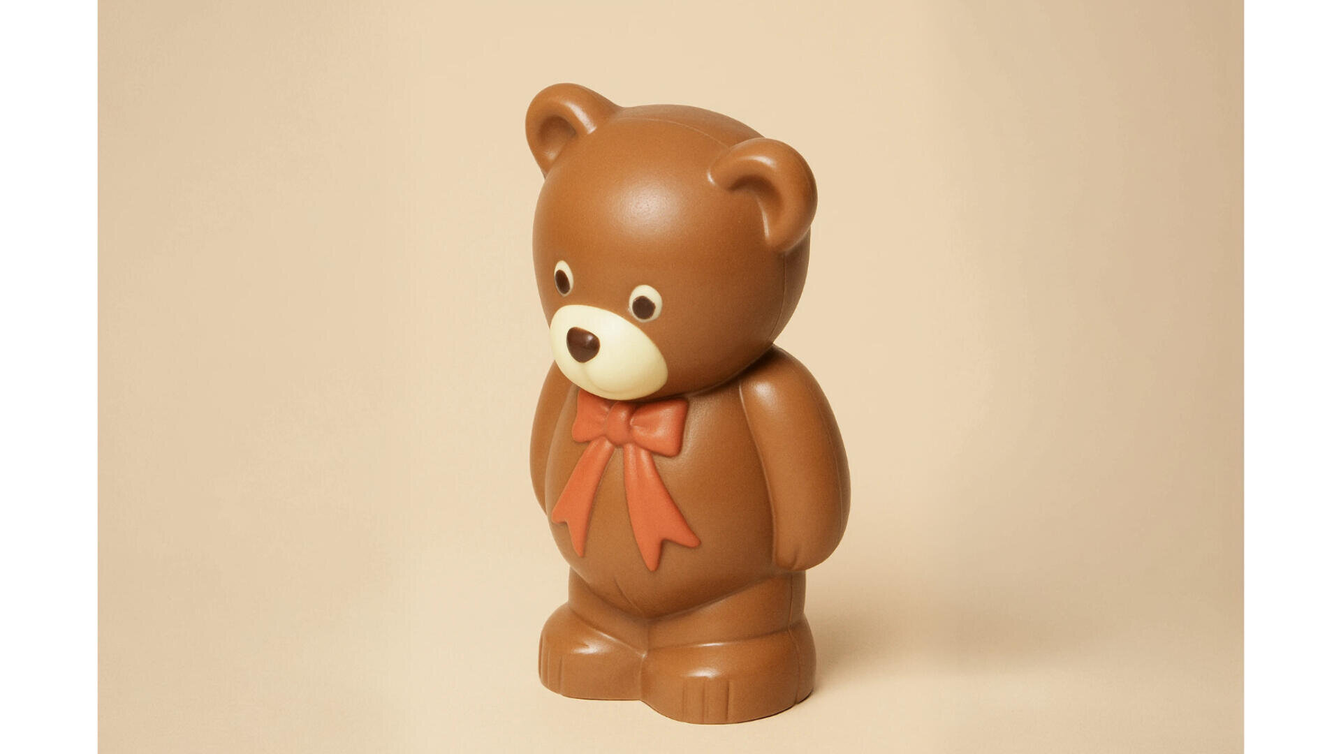 Vor einem schlichten beigen Hintergrund steht eine braune Teddybärfigur aus Kunststoff mit einer roten Schleife um den Hals.