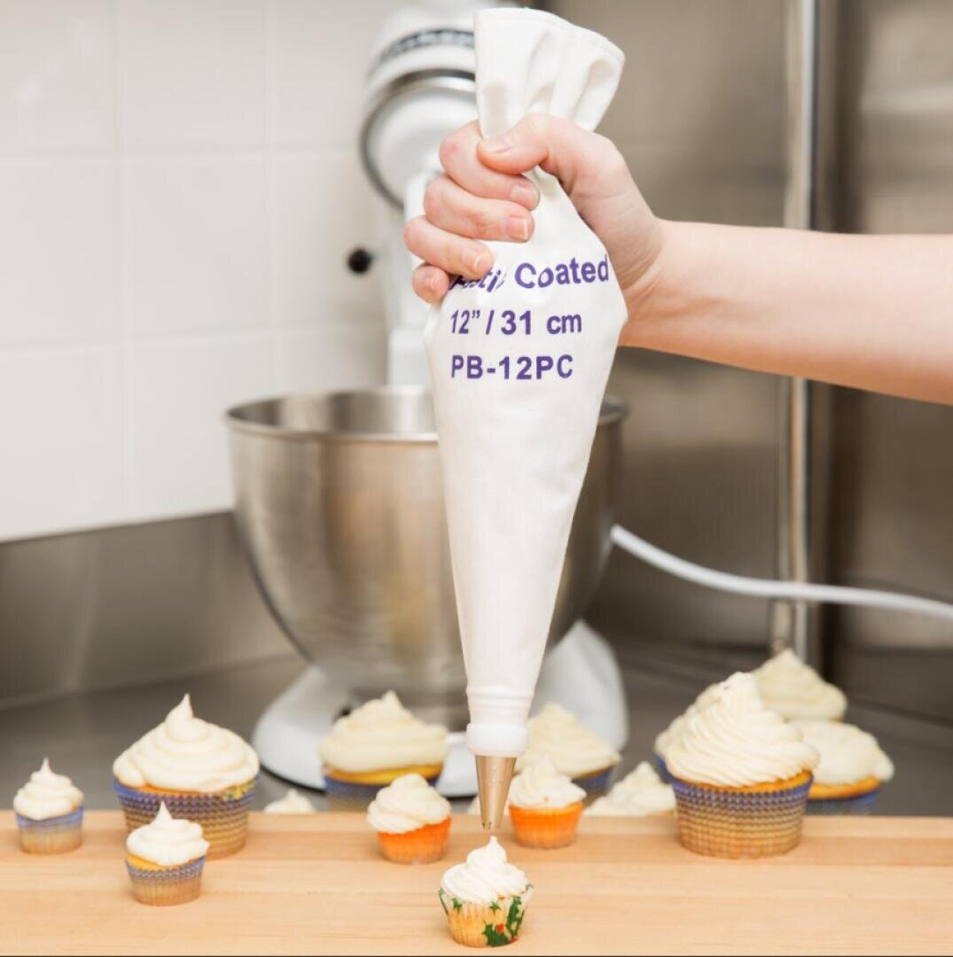 A hand is piping white frosting onto a cupcake with a piping bag. Several decorated cupcakes sit on a wooden surface, and a stand mixer is in the background.