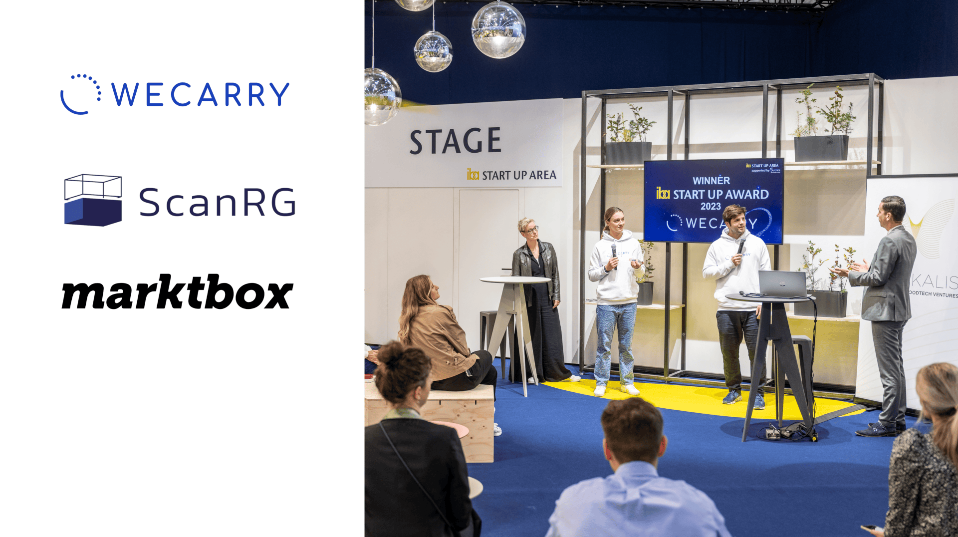People watch a startup award presentation on a stage where four individuals stand, two holding microphones. On the left, logos for WeCarry, ScanRG, and marktbox are displayed. The setting is modern and professional.