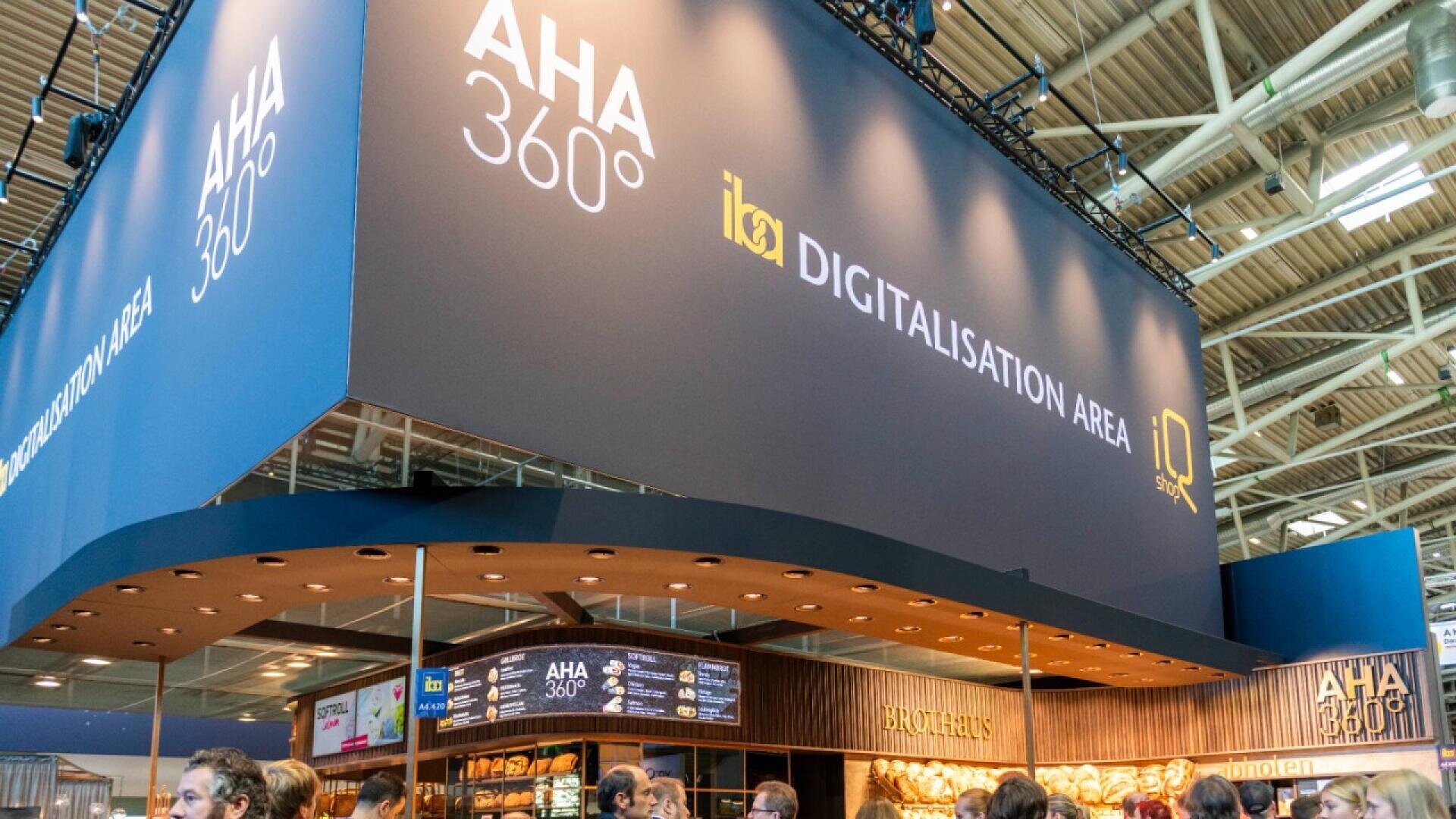 A large trade show booth displays signs reading AHA 360° and Digitalisation Area above a crowd of people. The booth has wooden accents and a section labeled Brotzeit. The setting is indoors with a high ceiling.