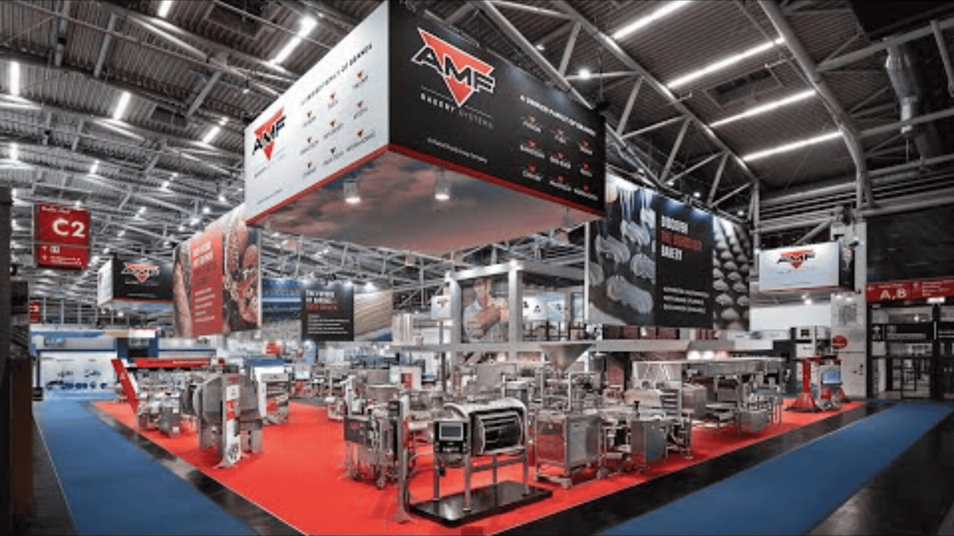 A large exhibition stand at a trade fair displays industrial bakery machines on a red carpet, with AMF branding overhead and information banners on the walls, surrounded by a spacious, modern hall.