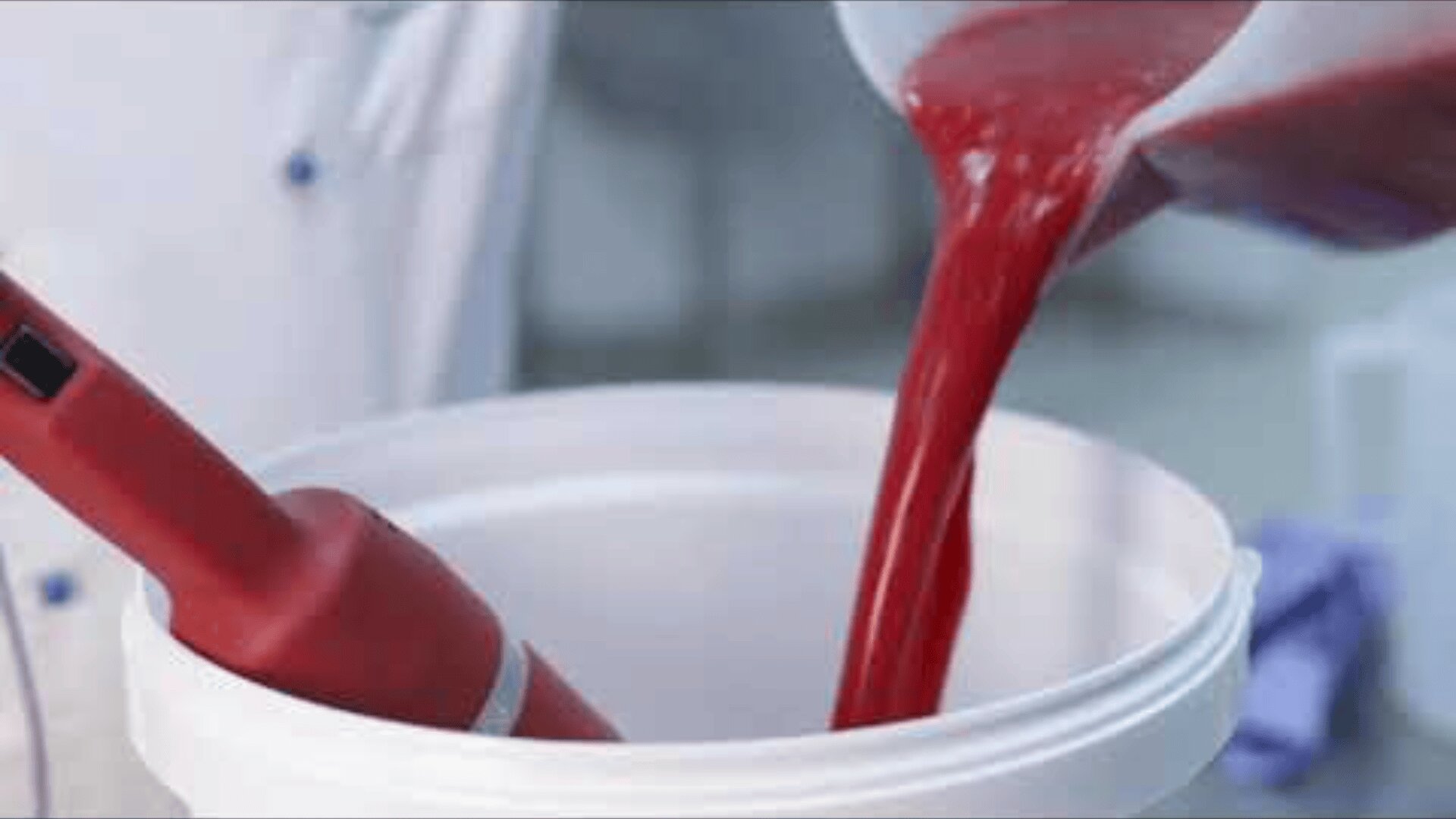 A thick red liquid is poured from a container into a white bucket with a red hand blender inside, probably in a laboratory or food processing facility.