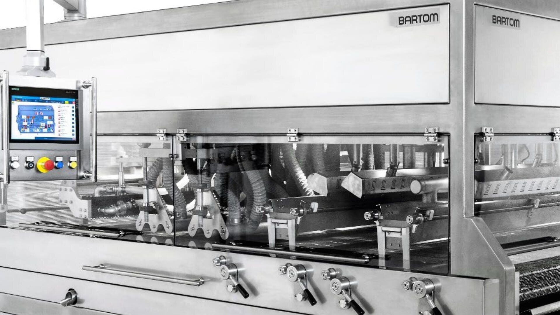 A stainless steel industrial food processing machine with a control panel displaying settings on the left; transparent panels reveal mechanical parts and conveyors inside. The brand name “Bartom” is visible on the machine.