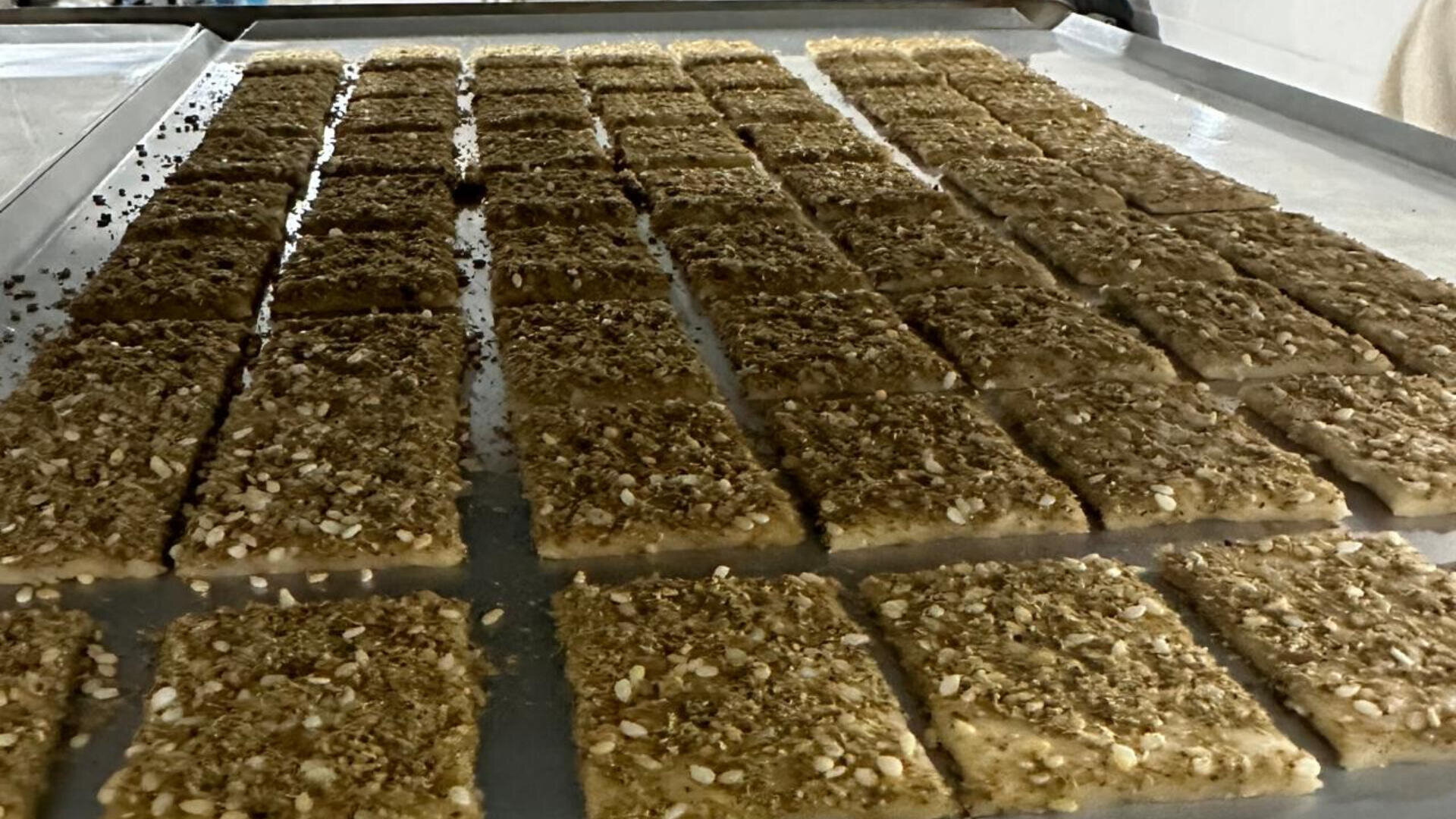 Rows of rectangular pastries topped with sesame seeds and spices are arranged on a large metal baking tray, ready to be baked or served.