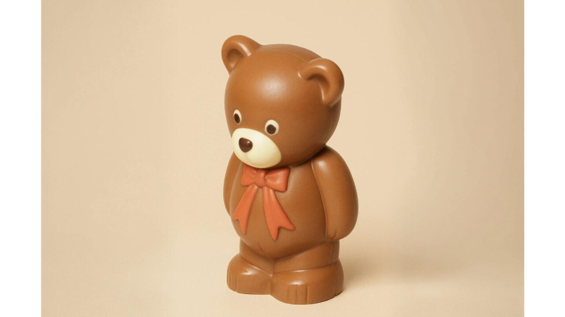 A brown teddy bear figurine with a red bow around its neck stands against a plain beige background. The bear has round ears, wide eyes, and a small, white snout.