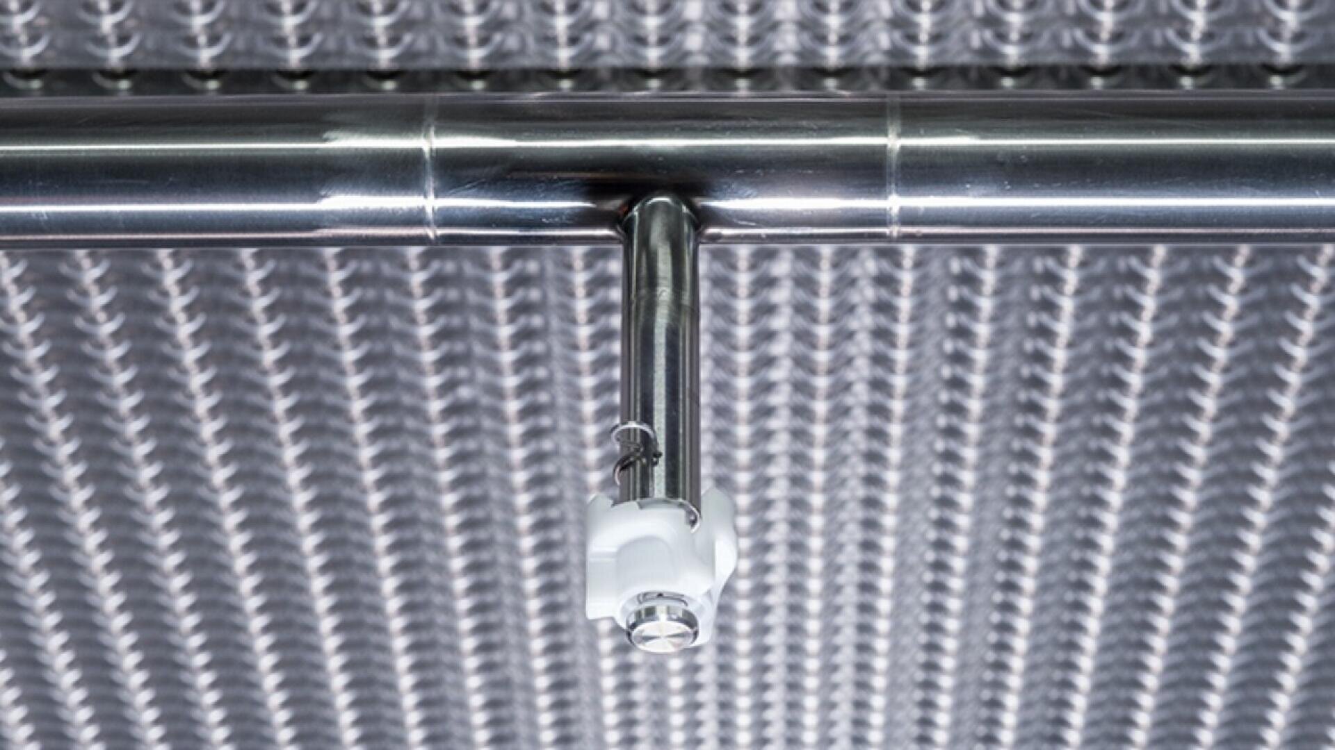 Nahaufnahme eines Sprinklerkopfs aus Edelstahl, der an einem Rohr befestigt und mit einem strukturierten, metallischen, gitterartigen Muster an einer Decke montiert ist.