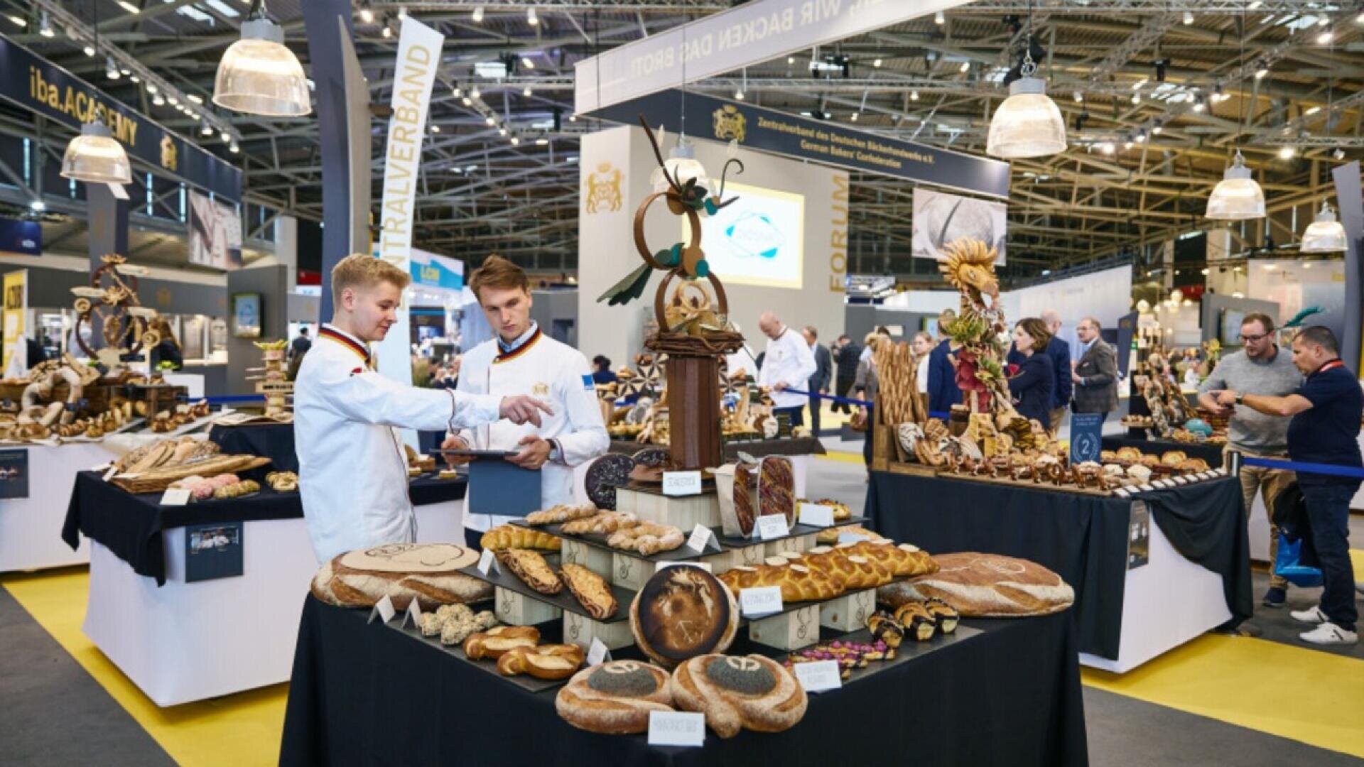 Two young men in white uniforms discuss near a display table filled with ornate bread and pastry creations at a large indoor exhibition, with more decorated tables and people in the background.