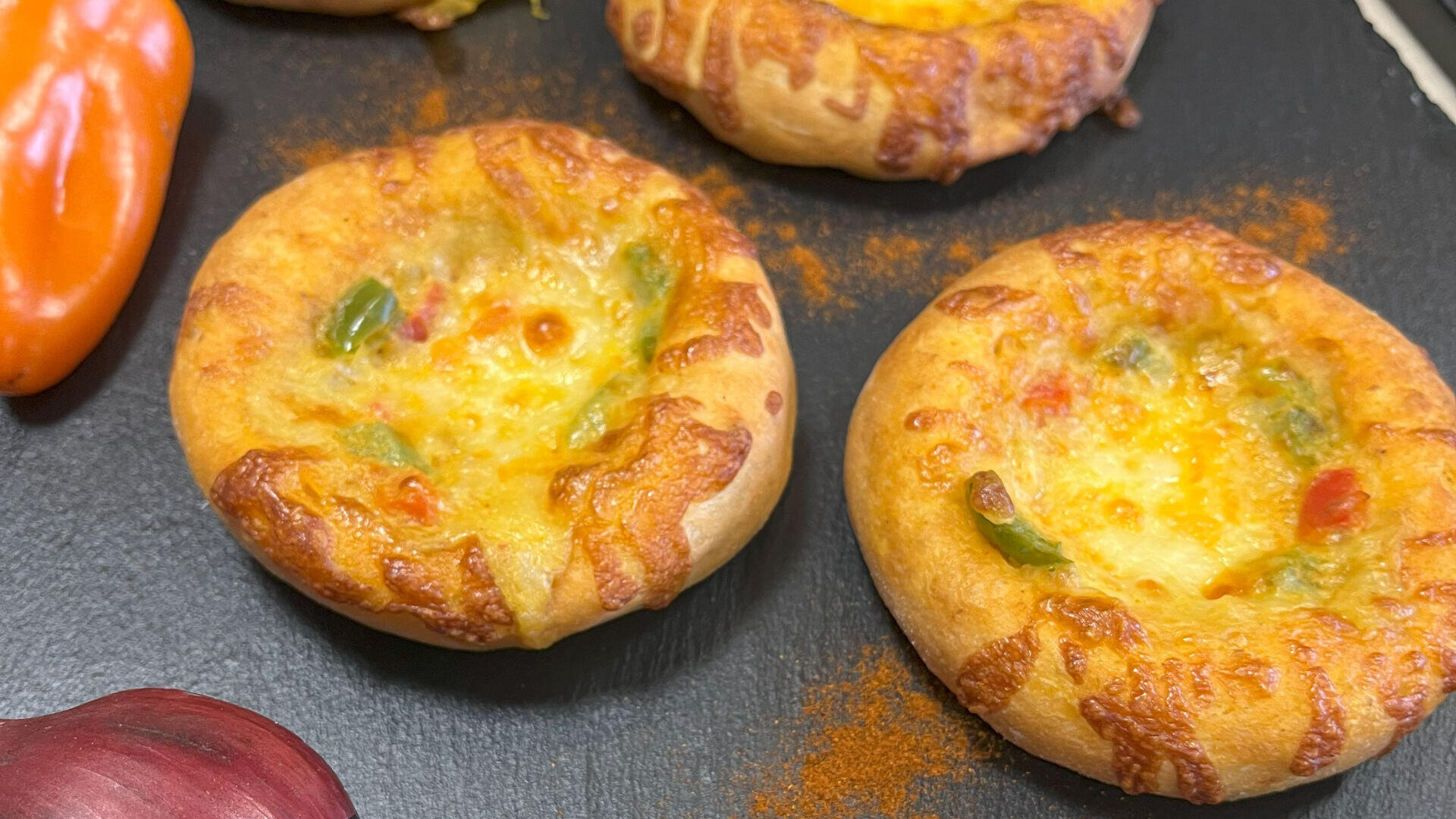 Zwei kleine, runde, gebackene Brotbecher, gefüllt mit geschmolzenem Käse und gewürfelten grünen und roten Paprikaschoten, stehen auf einer dunklen Oberfläche. In der Nähe sind auch eine rote Zwiebel und eine orange Paprikaschote teilweise zu sehen.