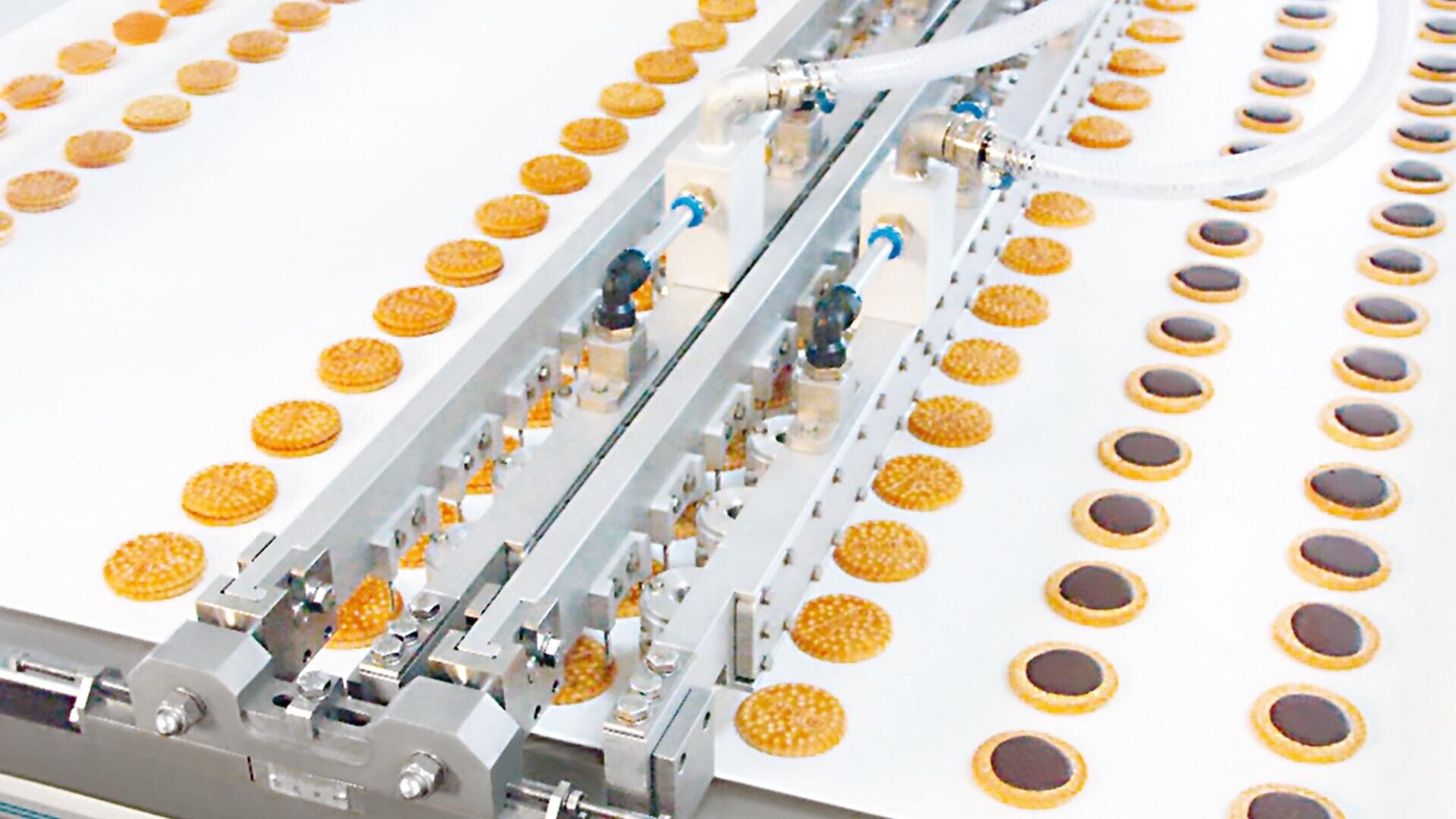 In einer Fabrik laufen Reihen von Keksen mit Schokoladenkern über ein Förderband, während eine automatische Maschine darüber Schokolade auf jeden einzelnen Keks verteilt.