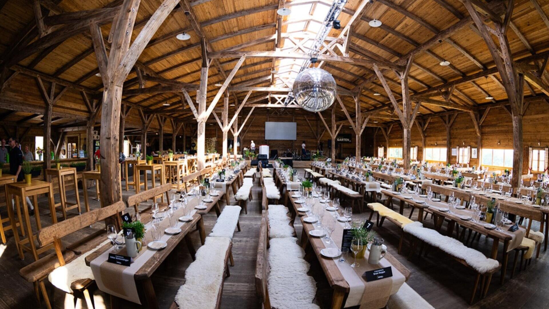 Large rustic wooden hall set up for an event, with long tables arranged in rows, neatly set with plates, glasses, and centerpieces. A large disco ball hangs from the ceiling, and natural light fills the space.