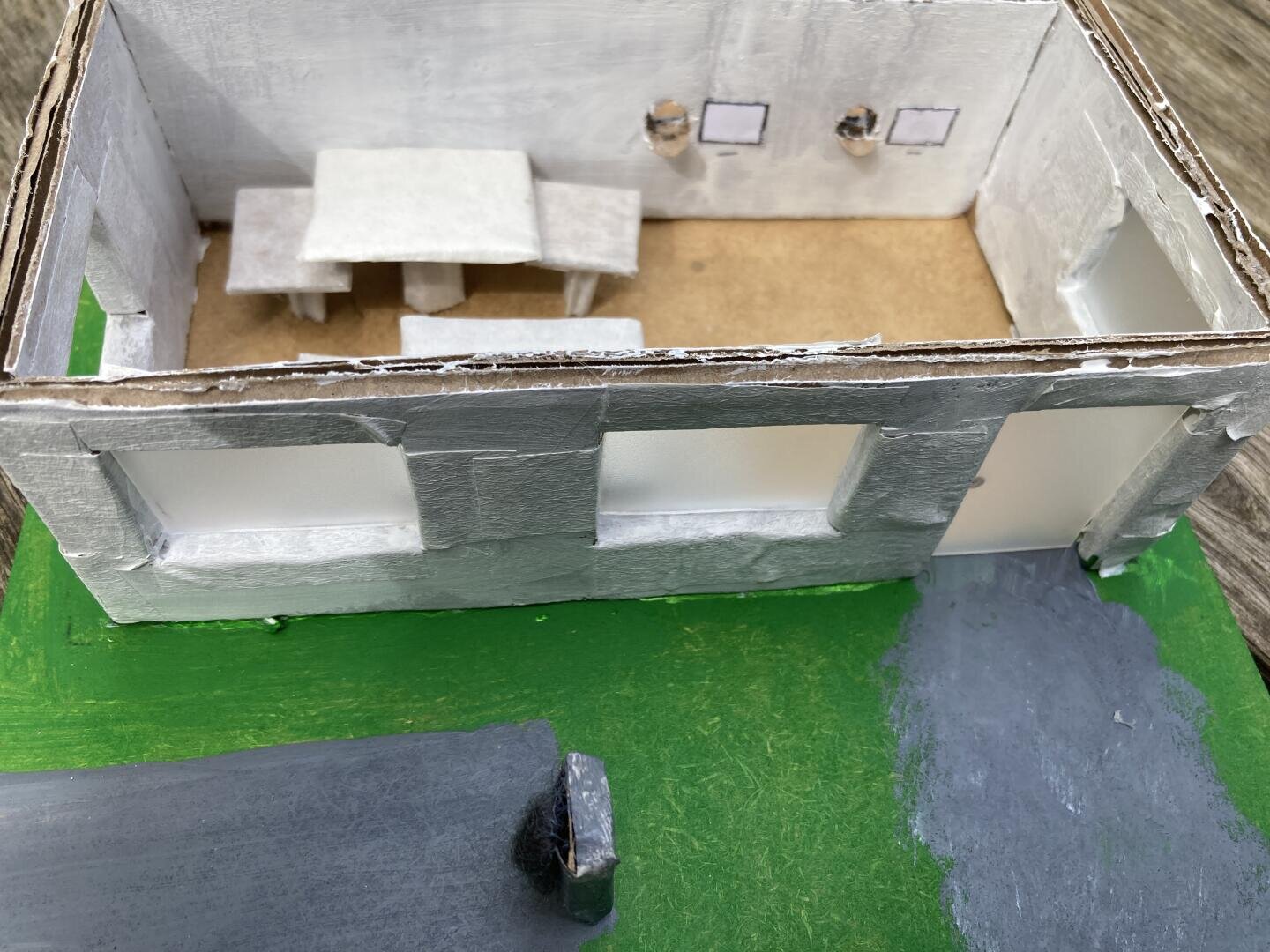 Ein handgefertigtes Modell eines Gebäudes mit weißen Wänden, großen Fenstern und grünem Sockel. Im Inneren befinden sich ein einfacher Tisch mit Bänken und zwei kleine gerahmte Gegenstände an der Wand. Die Szene besteht aus Pappe und Papier.