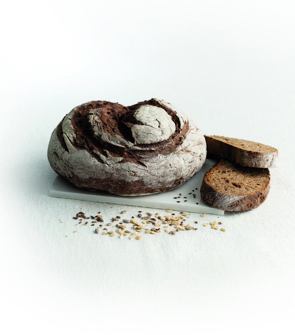 A round, rustic loaf of dark bread with a crusty exterior sits on a white surface alongside two slices and a small pile of seeds and grains scattered nearby.