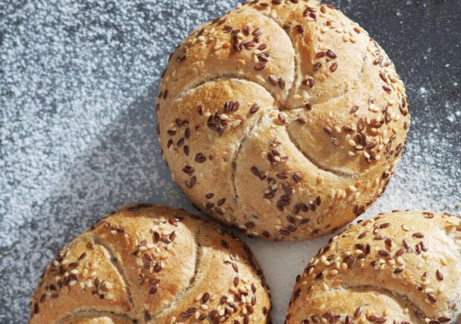 Three round, golden brown bread rolls with a swirl pattern and sprinkled with flax and sesame seeds rest on a lightly floured surface.