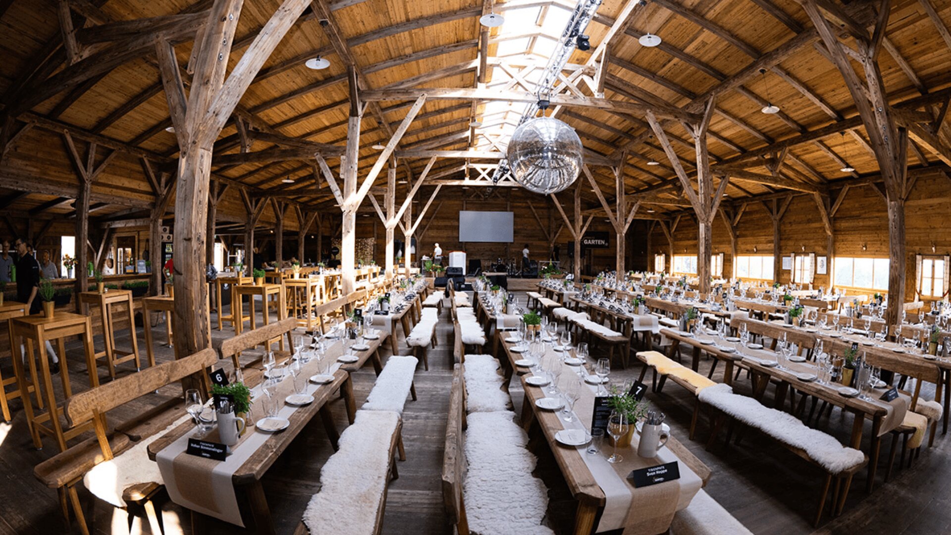 Eine große, rustikale Holzhalle, die für ein Bankett hergerichtet ist, mit langen Tischen und Bänken, die mit weißen Pelzdecken bedeckt sind, ordentlich angeordneten Tellern und Besteck, einer Discokugel, die von der Decke hängt, und Sonnenlicht, das durch Oberlichter fällt.