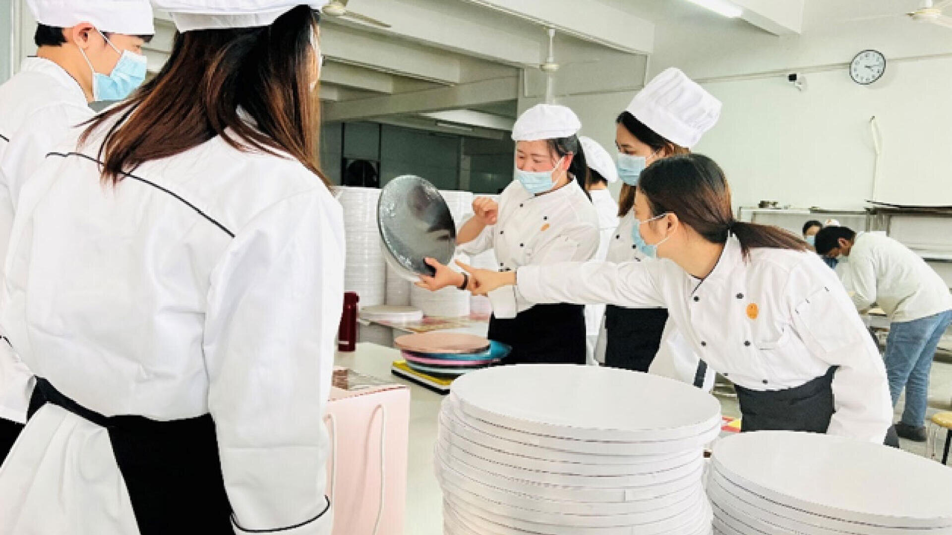 A group of people in white chef uniforms and face masks gather around a table in a kitchen, handling kitchenware and stacked white round trays. One person points while another holds a metal lid.