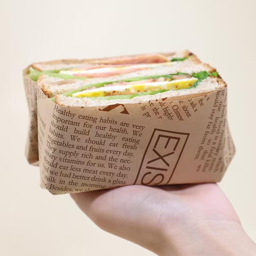 A hand holds a sandwich wrapped in newspaper-like paper; the sandwich contains lettuce, tomato, cheese, cucumber, and possibly ham, all between slices of whole wheat bread.