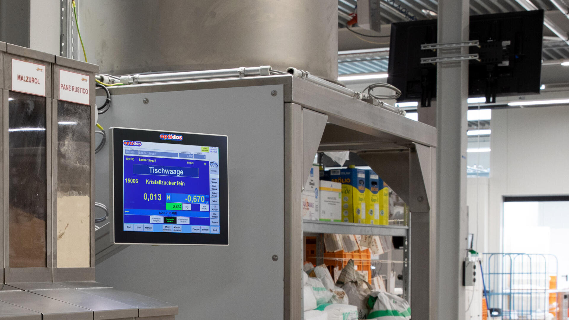 Eine an einer Industrieanlage in einem Lager angebrachte digitale Waage zeigt Gewichtsmessungen in Blau an. Im Hintergrund sind Regale mit verschiedenen Säcken und Behältern zu sehen.